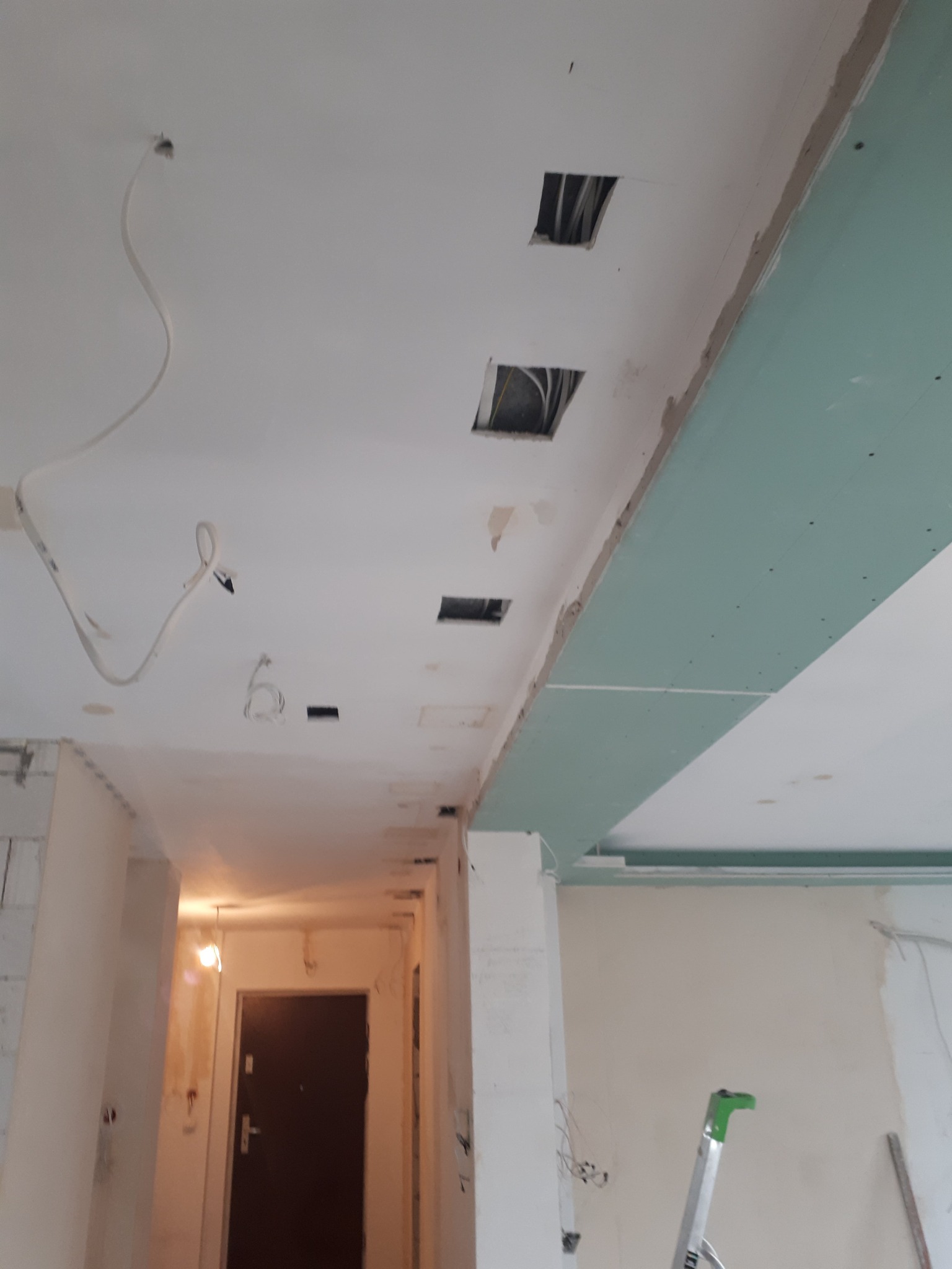 Fragment sufitu podwieszanego z widocznymi przewodami elektrycznymi w otworach, płyty gipsowo-kartonowe częściowo zamontowane, widoczna drabina w tle, wnętrze w trakcie remontu.