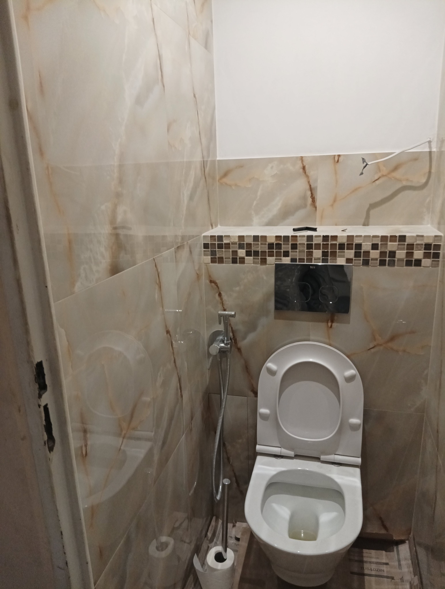 Wykończona toaleta z marmurowymi płytkami na ścianach, mozaiką dekoracyjną i wiszącą miską WC z bidetką.