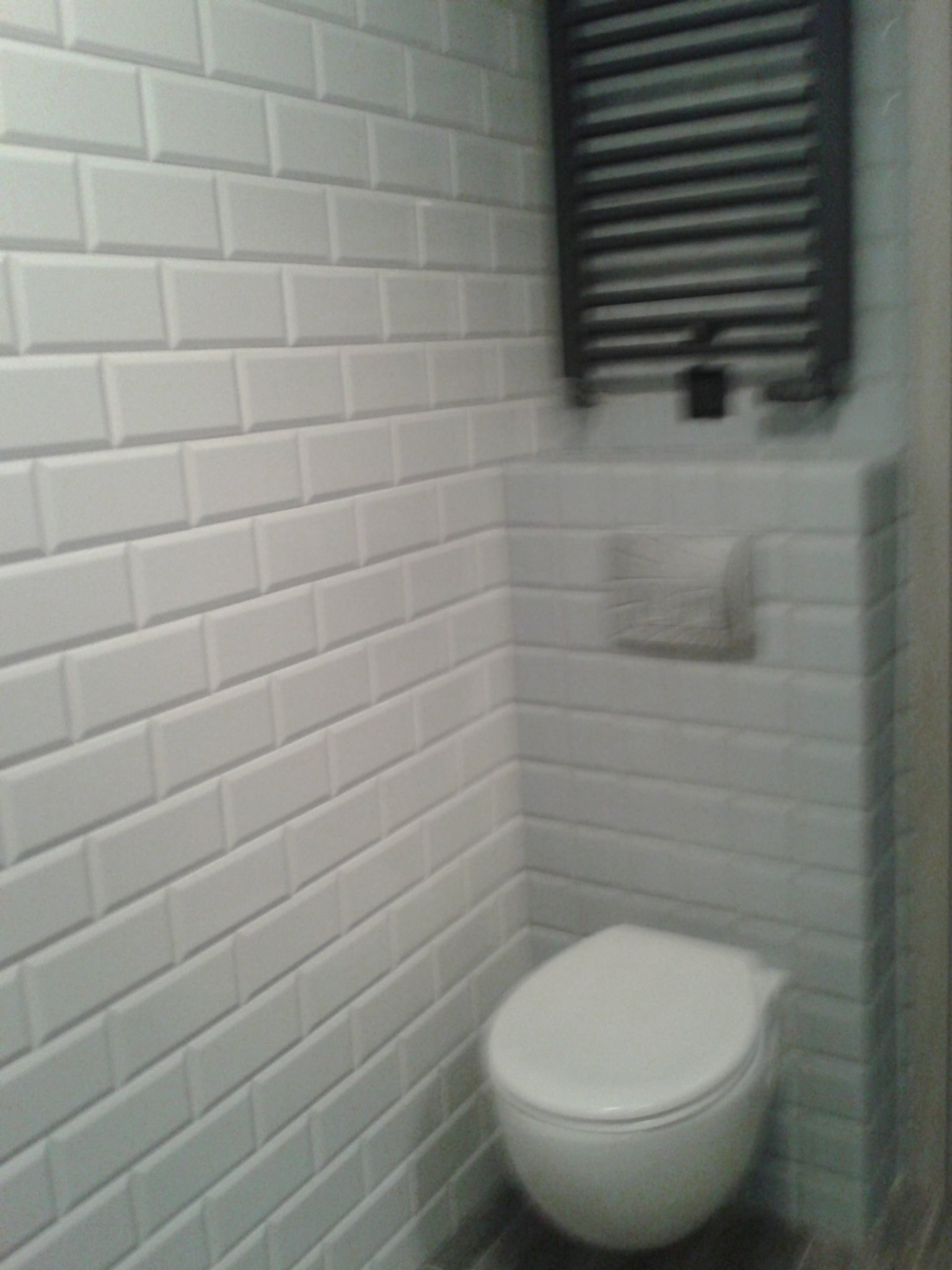 Wykończona łazienka z białą, prostokątną glazurą ułożoną w cegiełkę, wiszącą toaletą i grzejnikiem drabinkowym w kolorze grafitowym, zamontowanym nad zabudową geberitową.