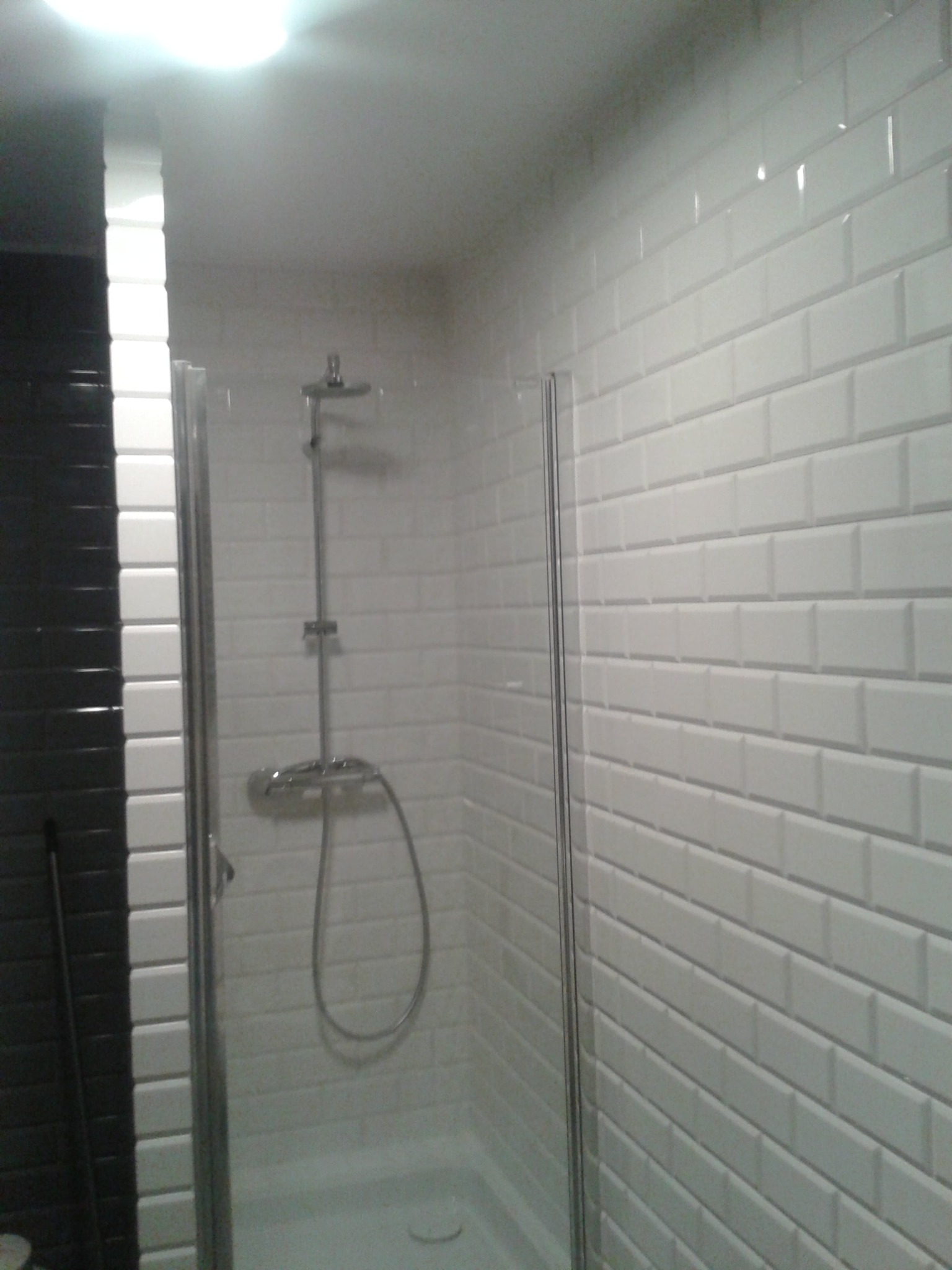 Kabina prysznicowa z białymi, prostokątnymi płytkami ułożonymi w stylu cegiełki, kontrastująca z ciemną ścianą obok, widoczny zestaw prysznicowy.