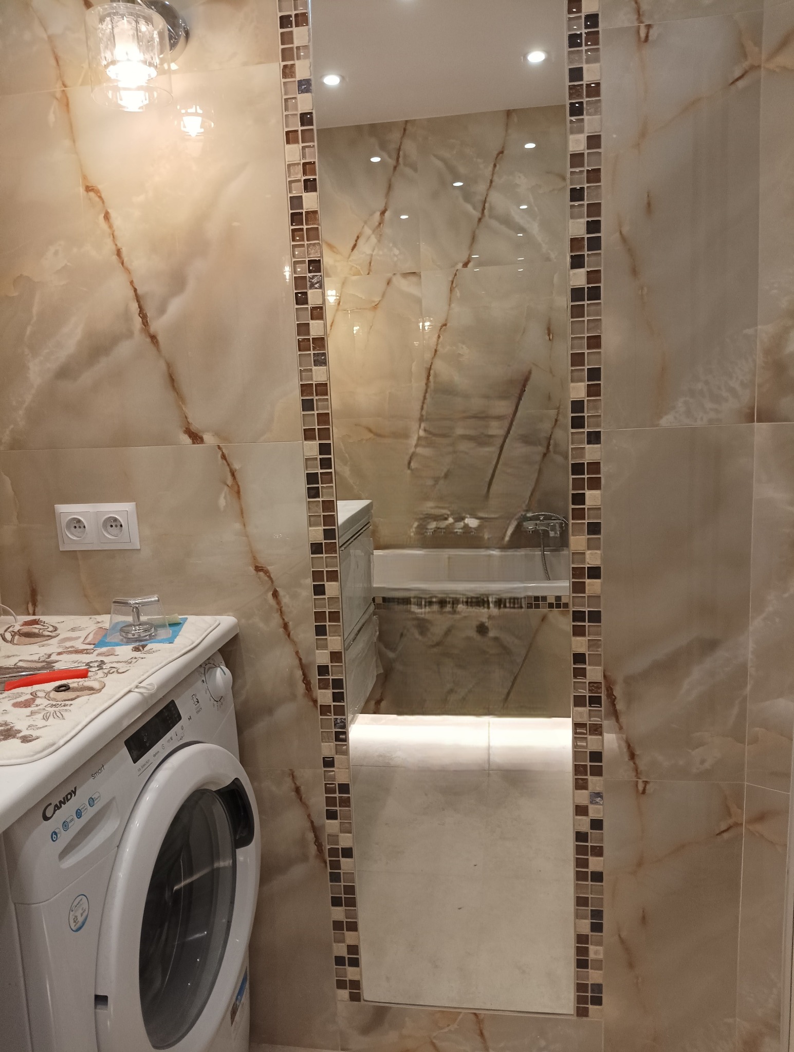Wykończona łazienka z beżowymi płytkami imitującymi marmur, lustro z mozaikową ramą odbija wannę, a obok stoi pralka Candy z akcesoriami na górze.