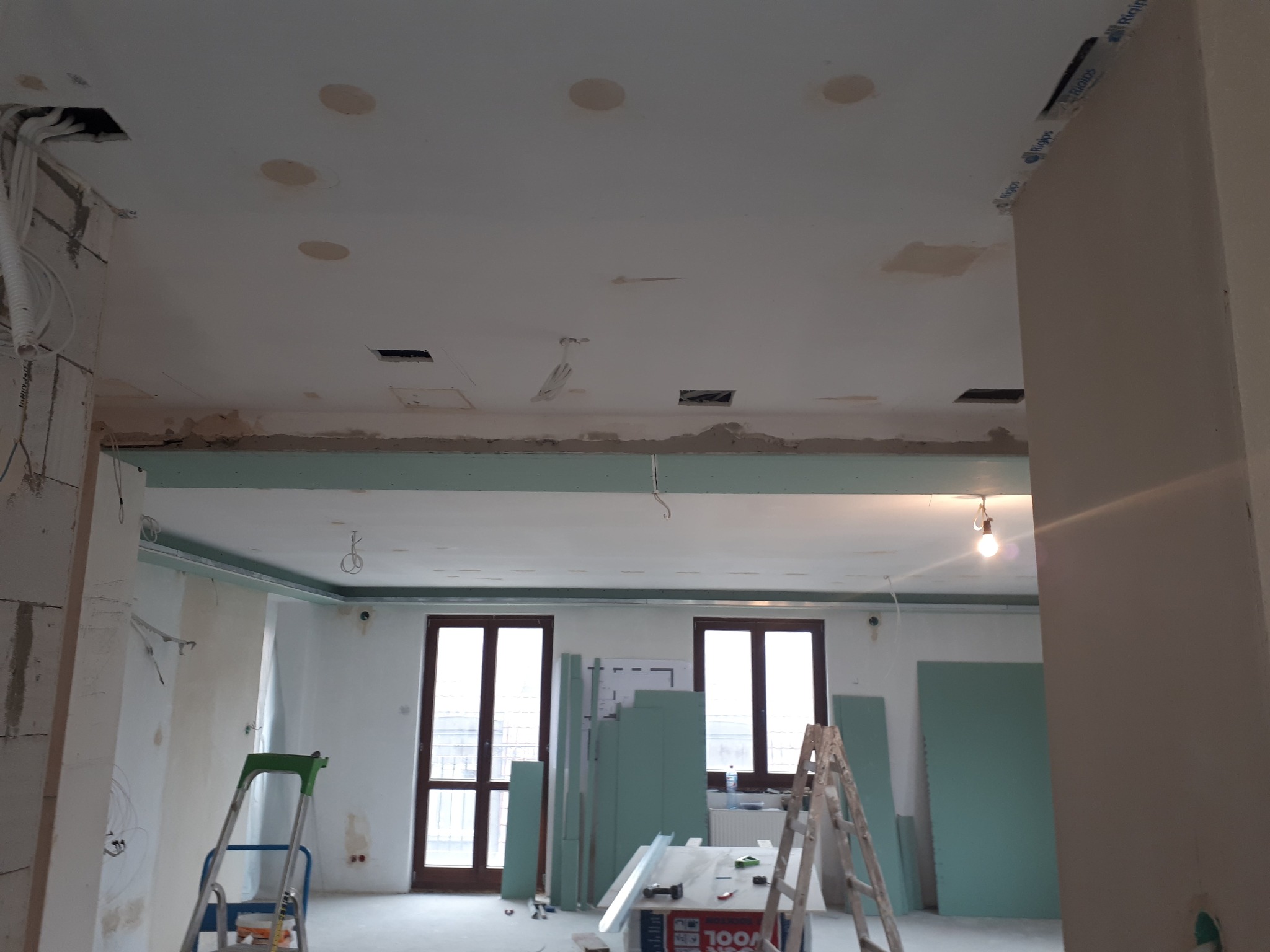 Wnętrze w trakcie remontu: widoczne płyty gipsowo-kartonowe, drabiny, kable wystające z sufitu, przygotowane otwory na oświetlenie punktowe oraz częściowo wykończone ściany.