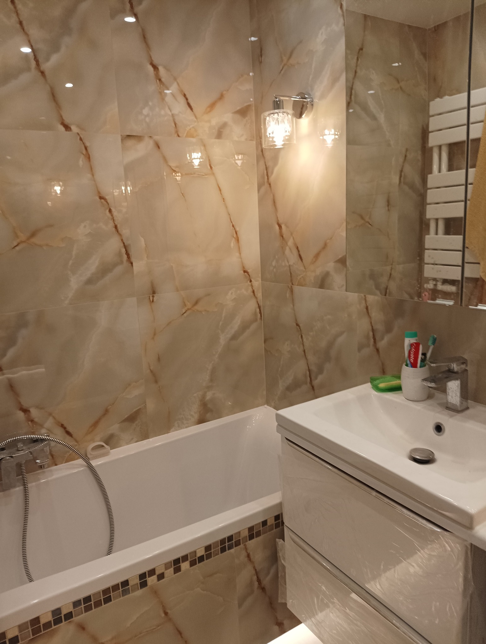 Wykończona łazienka z wanną obłożoną beżowymi płytkami imitującymi marmur z brązowymi żyłami, umywalką z szafką w folii ochronnej i dekoracyjną lampą ścienną.