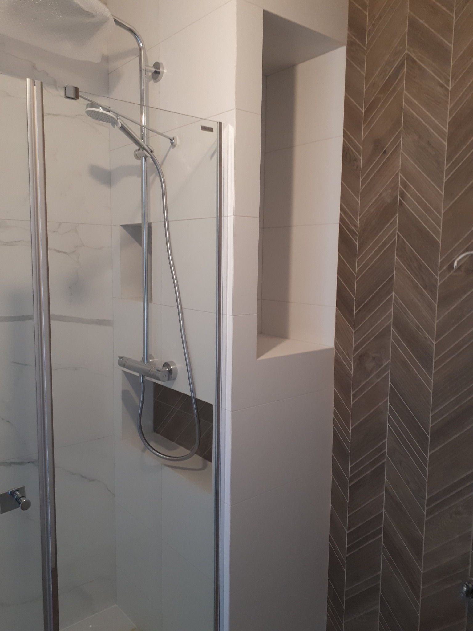 Nowoczesna kabina prysznicowa z chromowaną armaturą, białe płytki imitujące marmur i dekoracyjna ściana z płytek ułożonych w jodełkę, wnęka w ścianie.