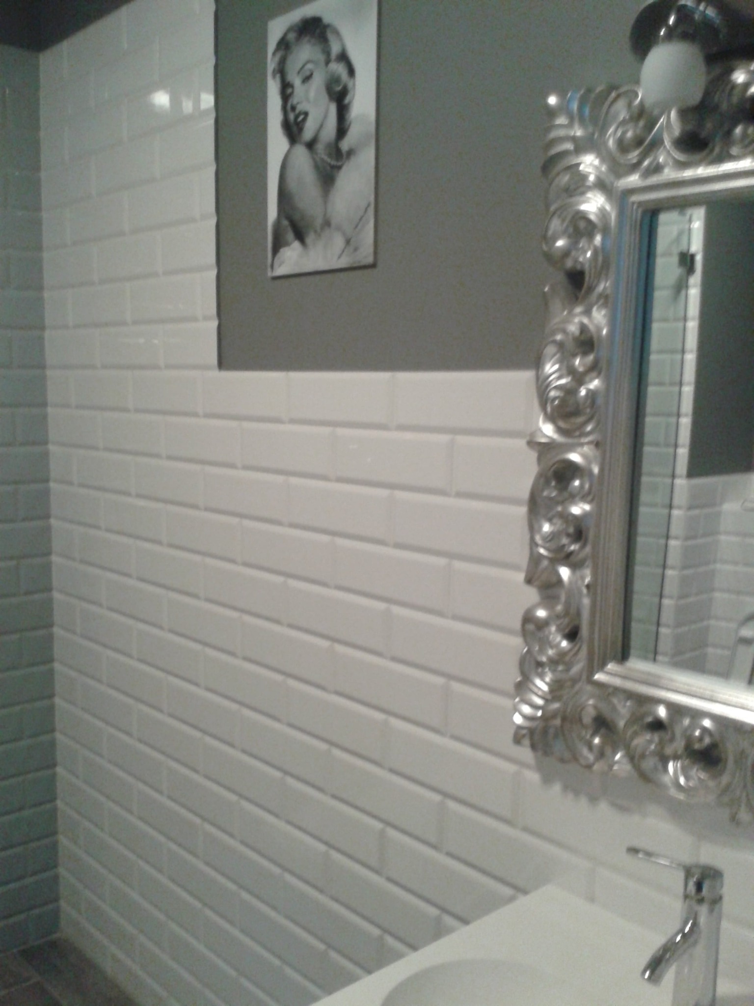Białe, prostokątne płytki ułożone w rzędach na ścianie łazienki, z lustrem w ozdobnej, srebrnej ramie i umywalką z baterią. Na ścianie wisi czarno-biały portret kobiety.