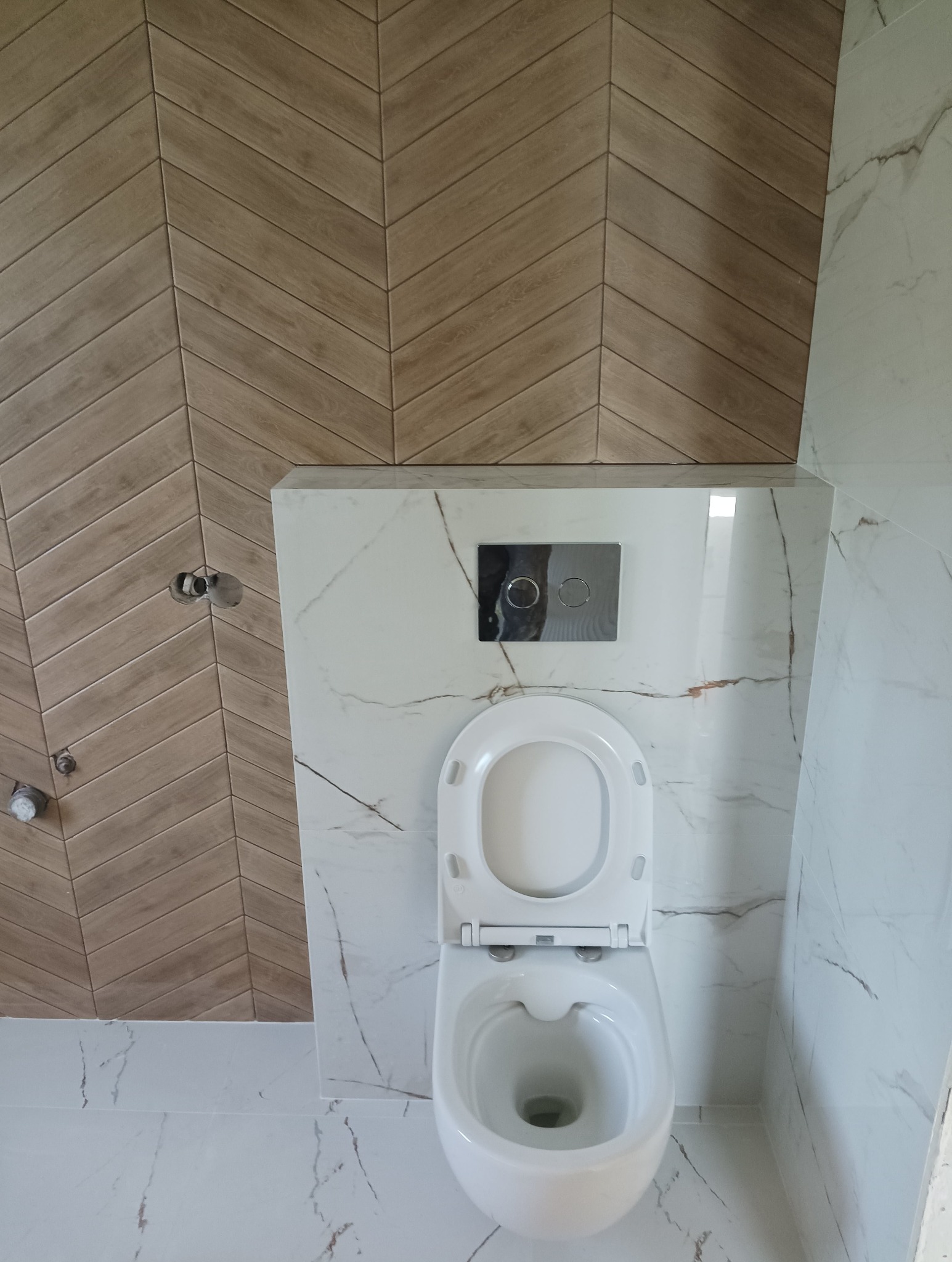 Nowoczesna toaleta podwieszana z otwartą klapą, marmurowe płytki na podłodze i ścianie, drewnopodobne płytki ułożone w jodełkę na ścianie, chromowany przycisk spłuczki.