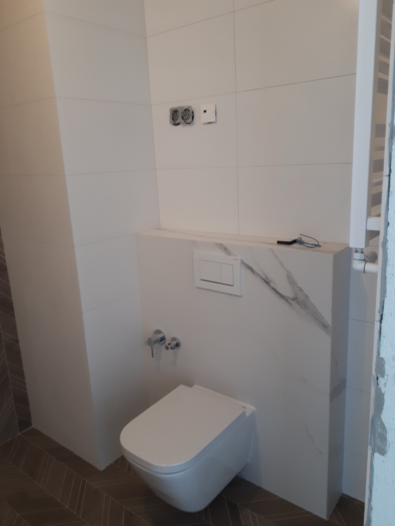 Nowoczesna toaleta podwieszana z białym przyciskiem spłukującym, umieszczona na ścianie wykończonej płytami imitującymi marmur, z widocznymi chromowanymi zaworami i częściowo widocznym grzejnikiem...