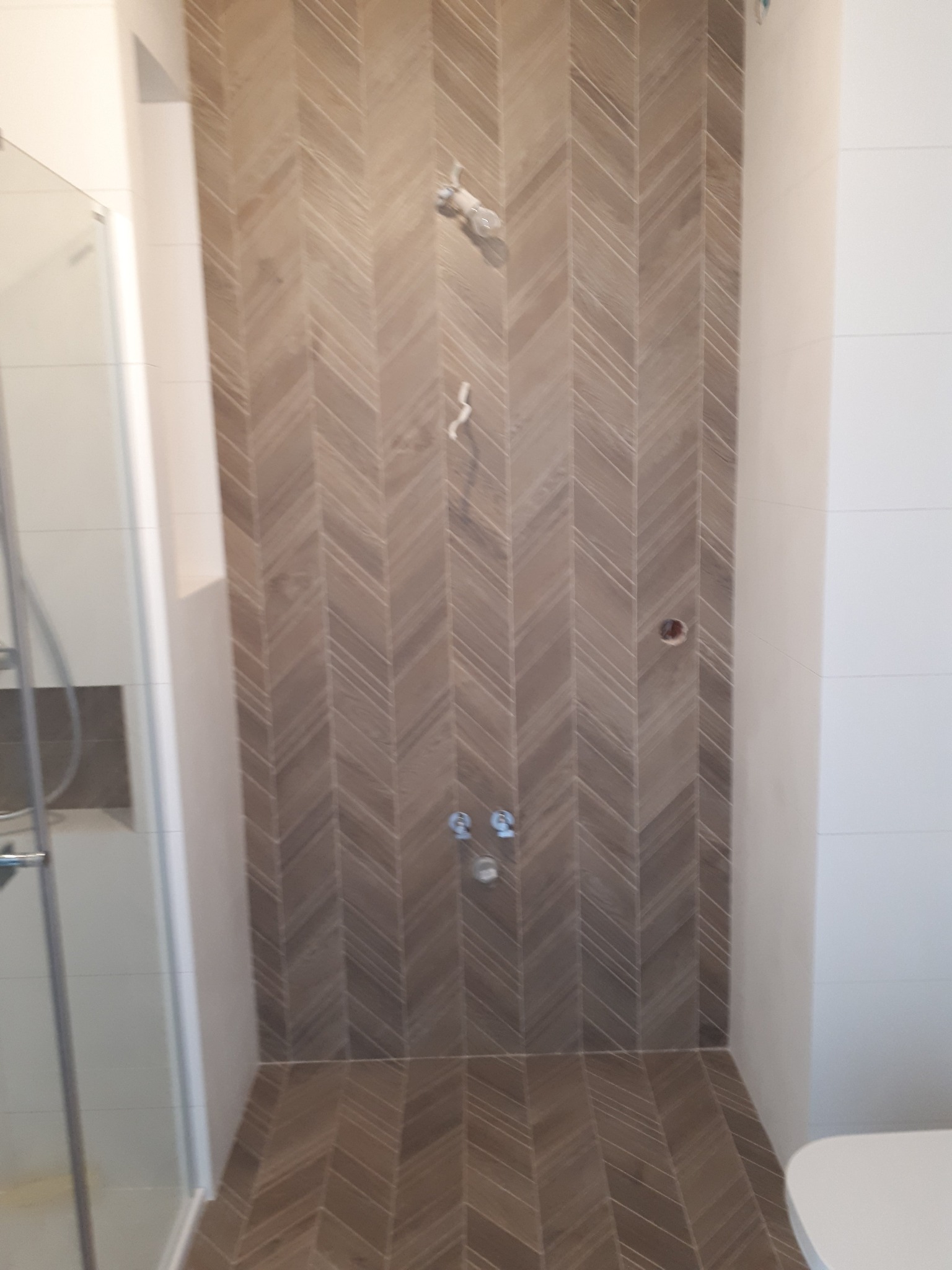 Ściana i podłoga w kabinie prysznicowej wyłożone brązowymi płytkami w jodełkę, widoczne przygotowane punkty instalacyjne, białe ściany boczne.