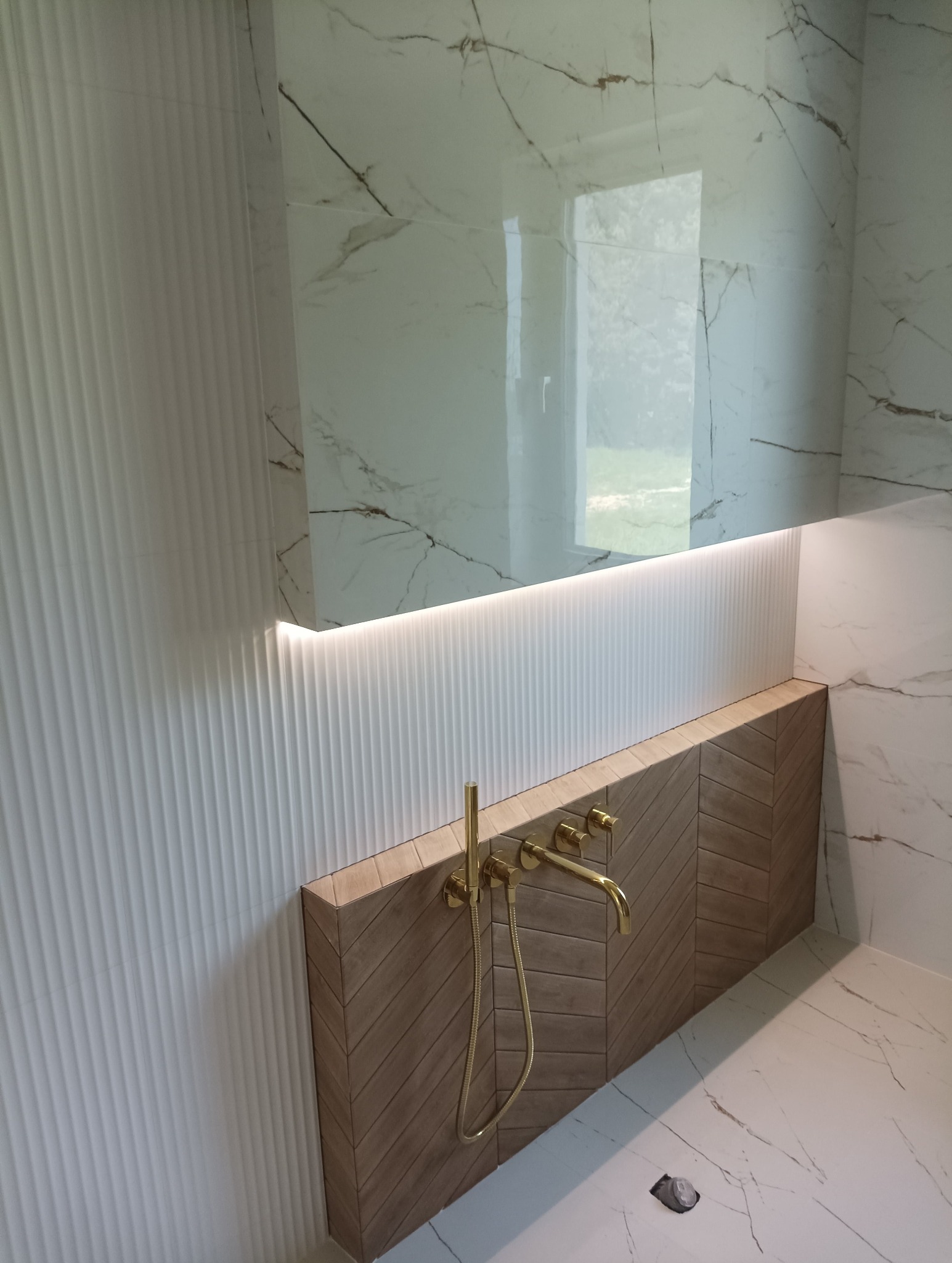 Nowoczesna łazienka z białymi, marmurkowymi płytkami, strukturalną ścianą i drewnopodobną obudową w jodełkę z zamontowaną złotą armaturą.