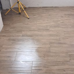 S&M HOUSE MAKSYM SHAPOVAL - Ułożone, podłużne płytki podłogowe imitujące drewno, oświetlone lampą budowlaną. Widoczny fragment ściany w trakcie prac wykończeniowych.