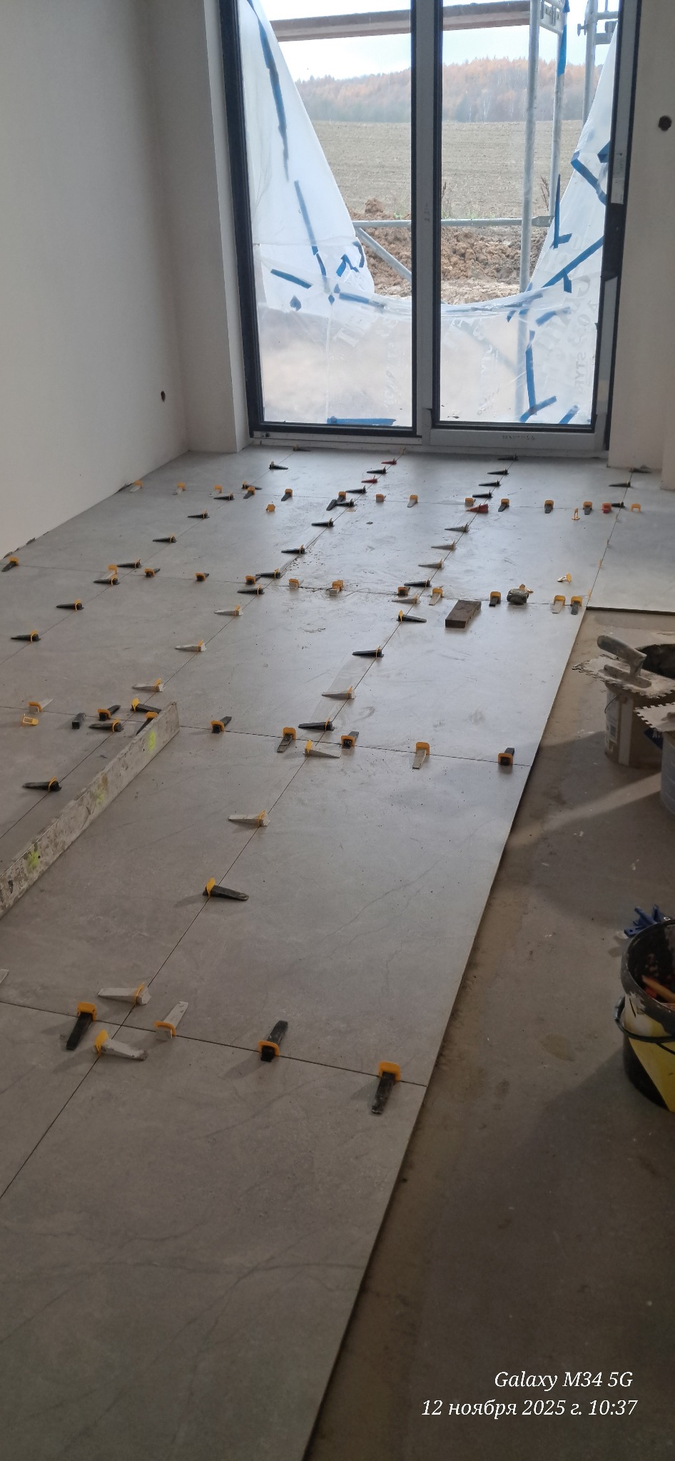 Układanie płytek podłogowych w jasnym wnętrzu, z użyciem klipsów poziomujących. Widoczne narzędzia i okno z folią ochronną. Praca w toku, surowy stan.