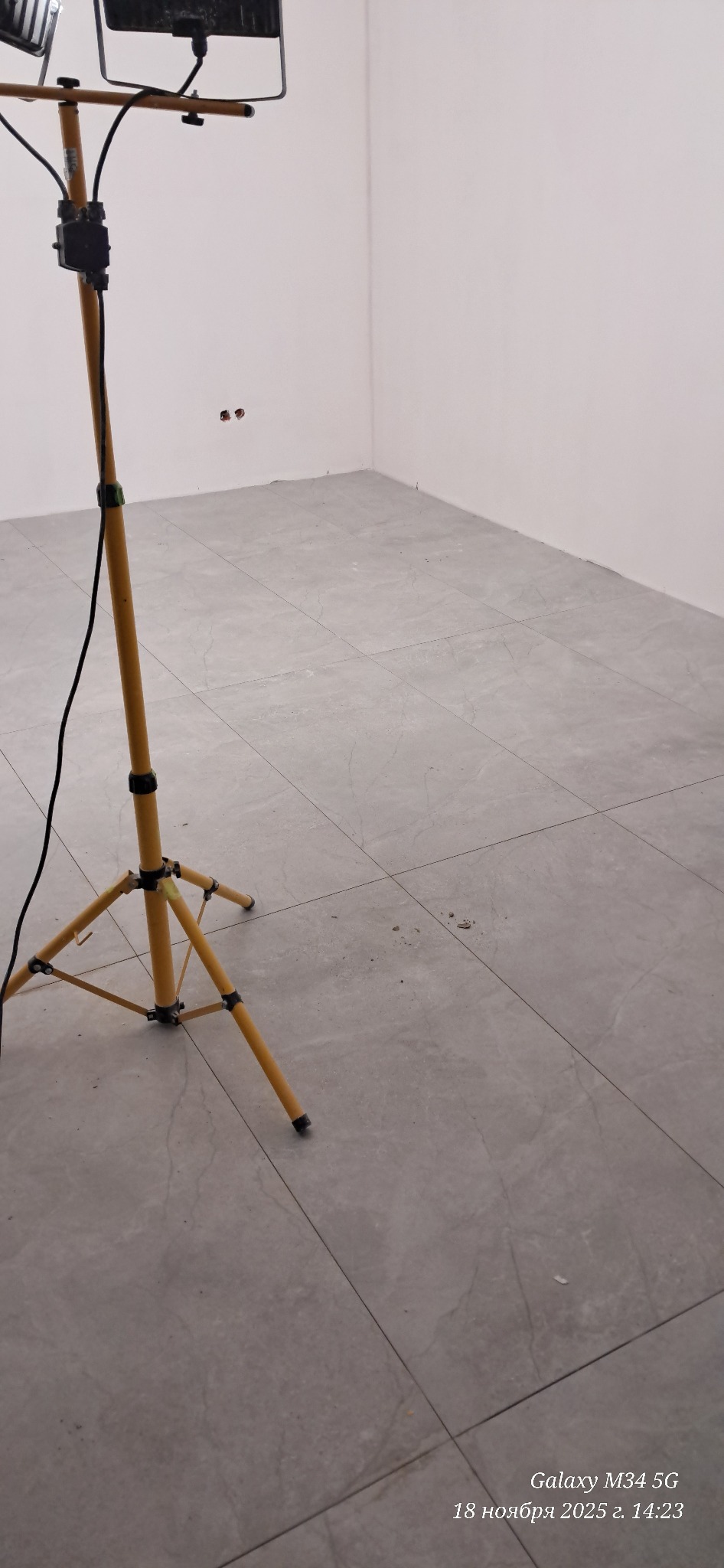 Szare płytki podłogowe w rogu pomieszczenia oświetlone lampą budowlaną. Widoczne gniazdka elektryczne na ścianie. Minimalistyczny design wnętrza po remoncie.