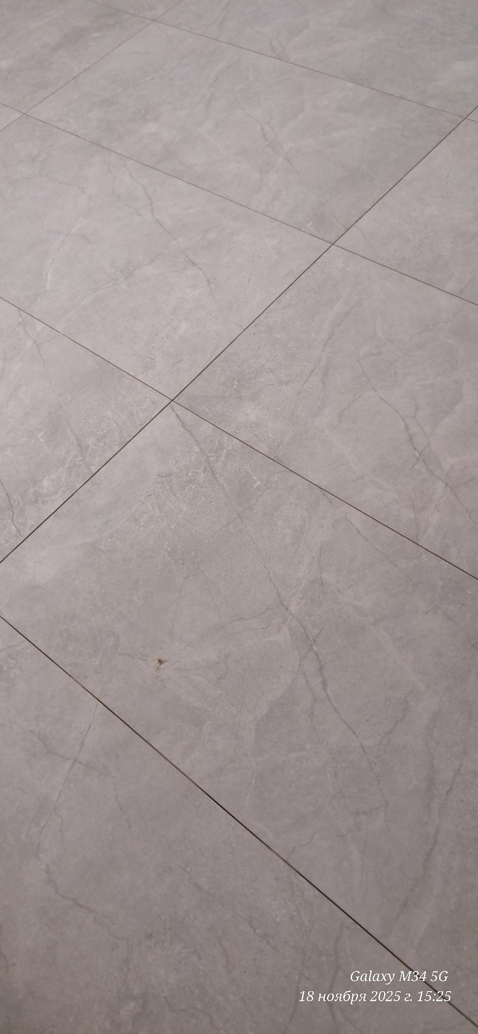 Szare płytki podłogowe z delikatnym wzorem marmuru, ułożone ukośnie. Widoczna fuga i drobne zabrudzenie na jednej z płytek. Zdjęcie z bliska.