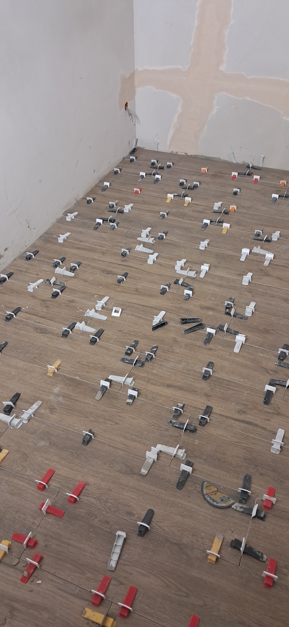 Układanie płytek podłogowych imitujących drewno z użyciem systemu poziomowania. Widoczne klipsy i kliny. Ściana przygotowana do gładzi.