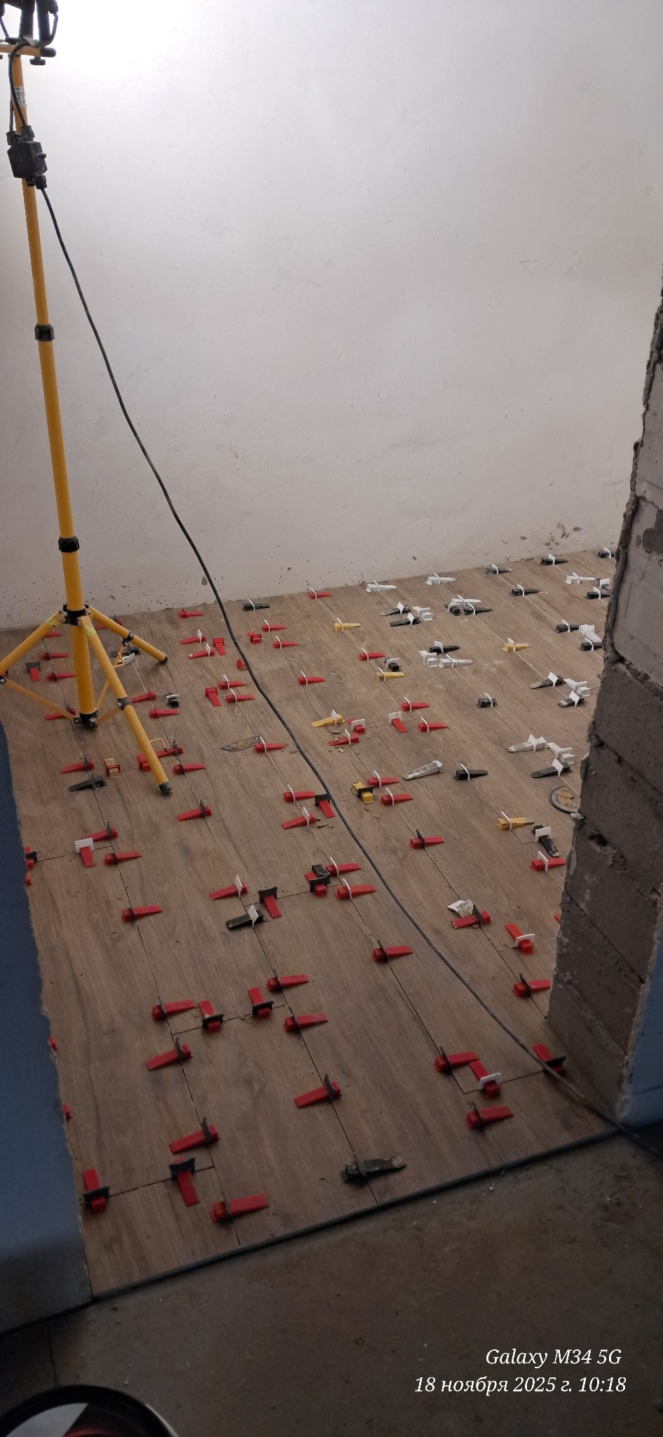 Układanie płytek podłogowych z użyciem klipsów poziomujących, oświetlone lampą budowlaną. Widoczne fragmenty ścian i surowego muru.