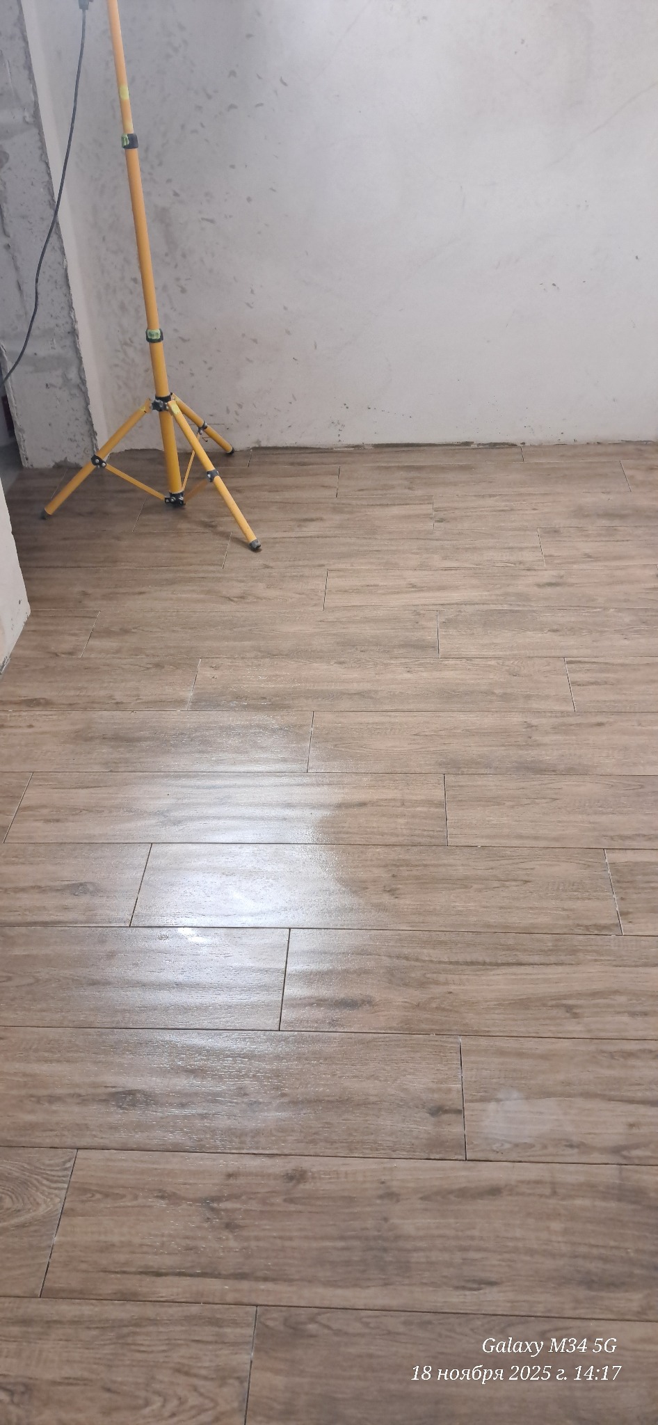 Ułożone, podłużne płytki podłogowe imitujące drewno, oświetlone lampą budowlaną. Widoczny fragment ściany w trakcie prac wykończeniowych.