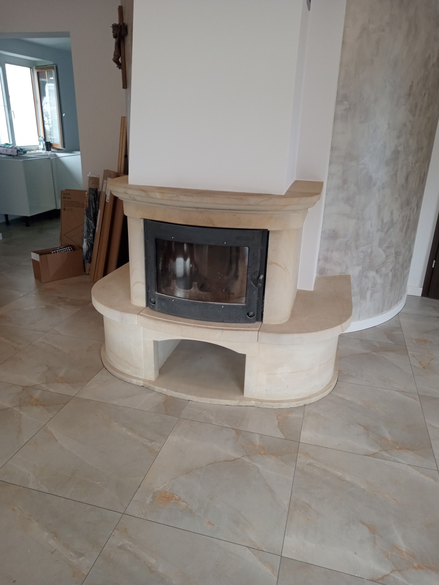 Jasny, kamienny kominek z czarnym wkładem kominkowym, widoczny na tle jasnych płytek podłogowych i ściany z efektem betonu.