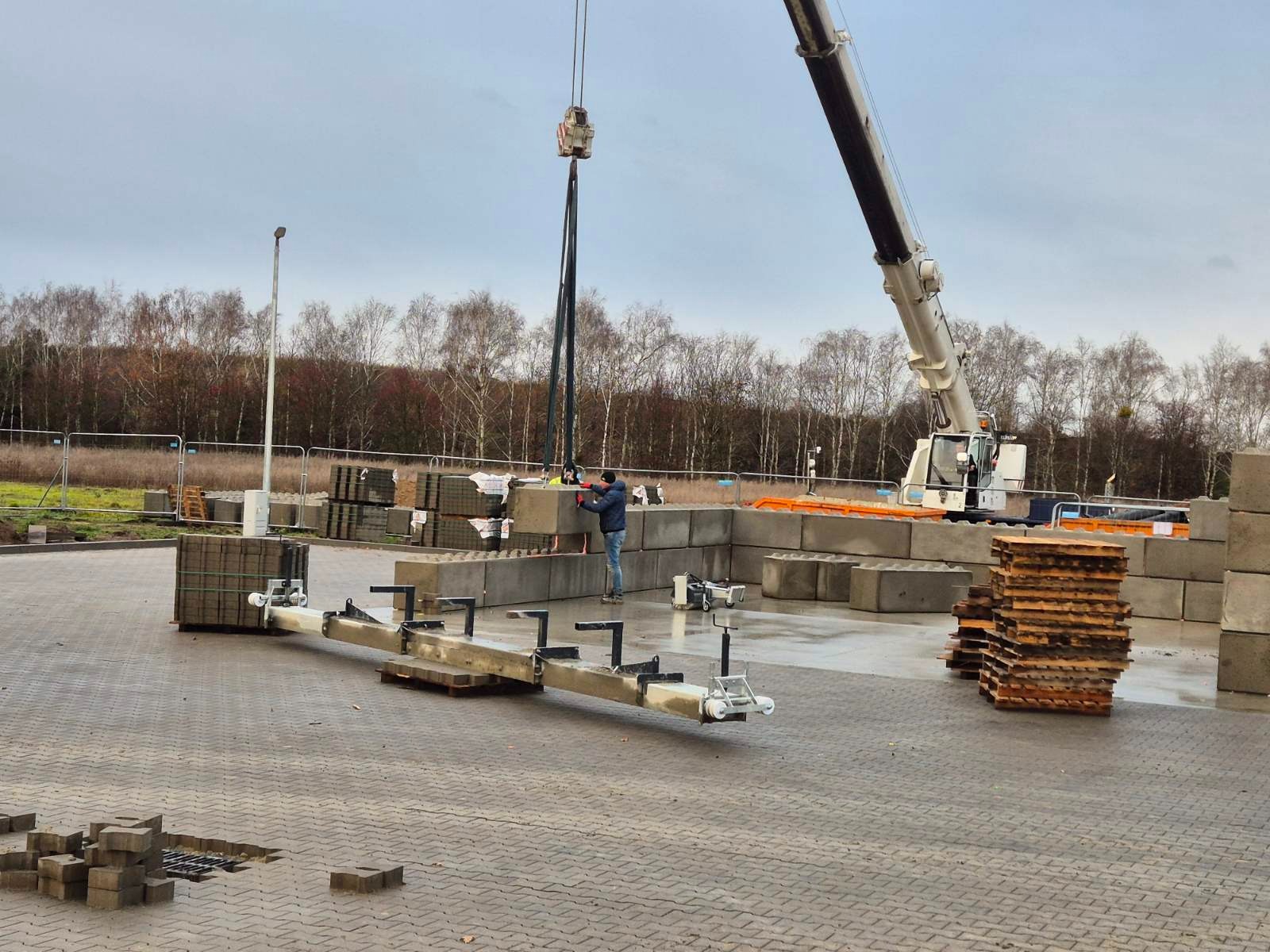Montaż prefabrykowanych betonowych bloków za pomocą dźwigu na utwardzonym placu, widoczny pracownik w odzieży roboczej, palety z kostką brukową w tle.