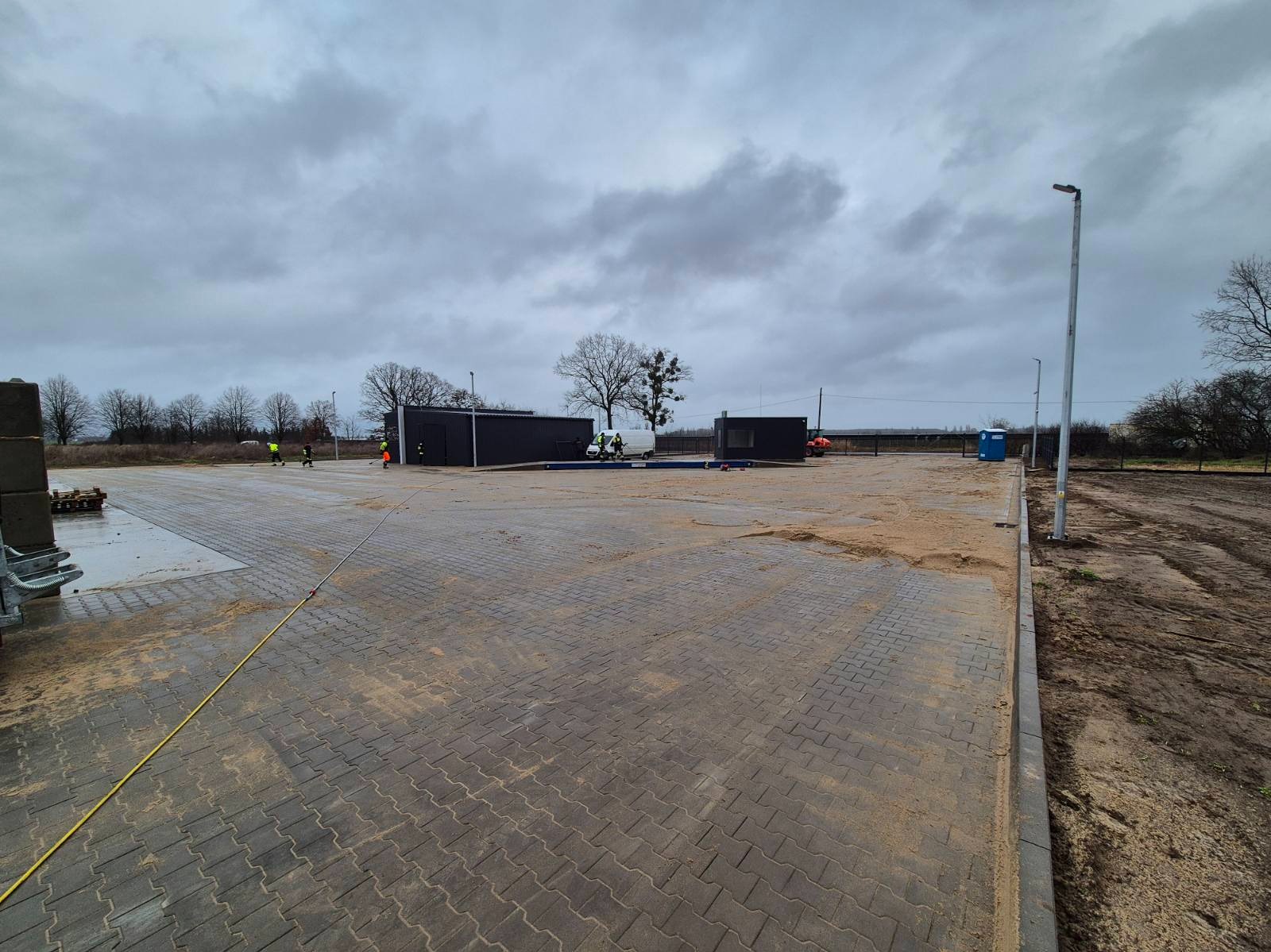 Ułożona szara kostka brukowa na dużym placu, z widocznymi czarnymi kontenerami biurowymi, pracownikami w odblaskowych kamizelkach i rozciągniętym żółtym kablem, całość sfotografowana w pochmurny...