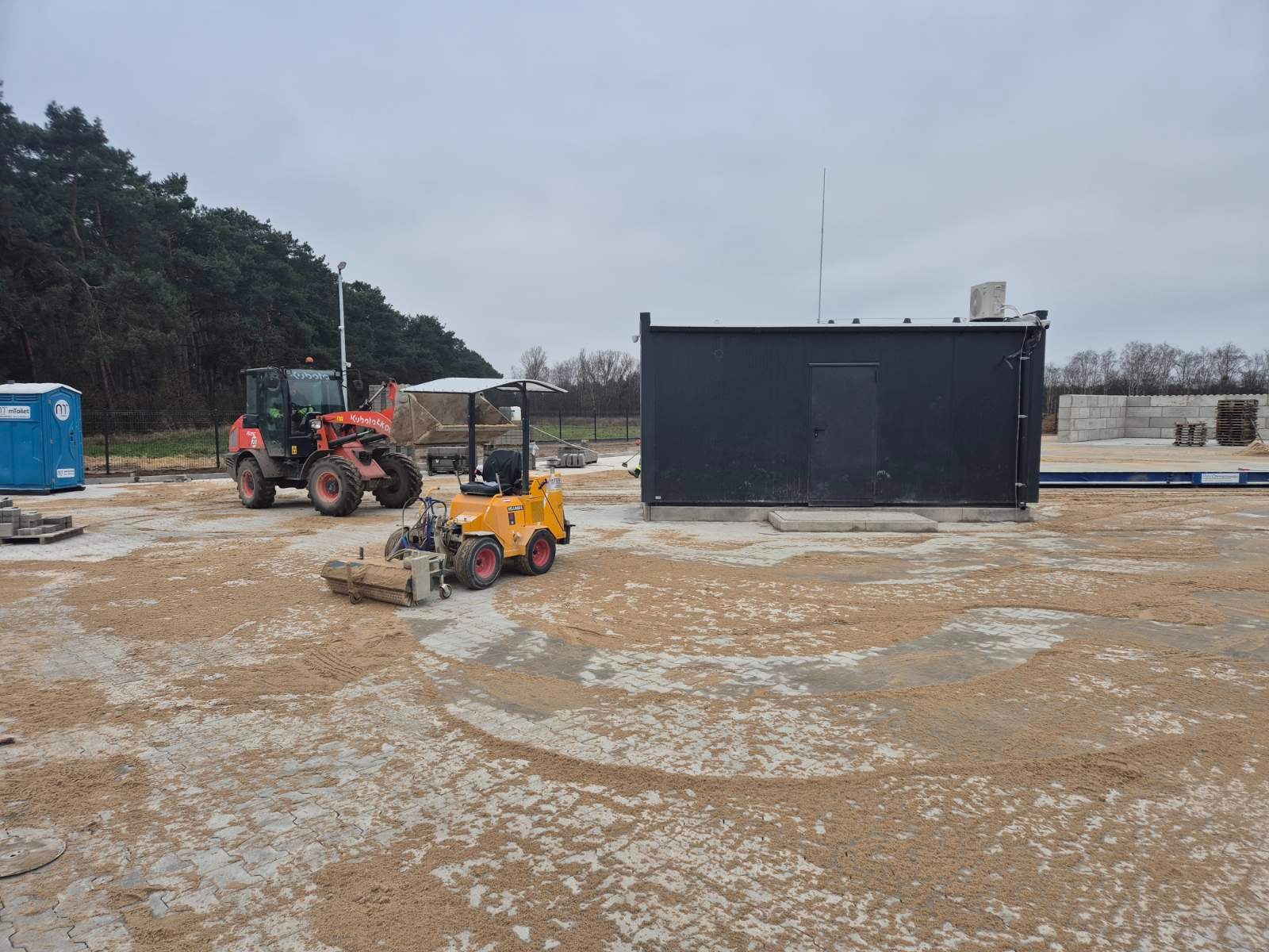 Układanie kostki brukowej z użyciem mini zamiatarki, widoczny także wózek widłowy i przenośna toaleta na placu budowy.
