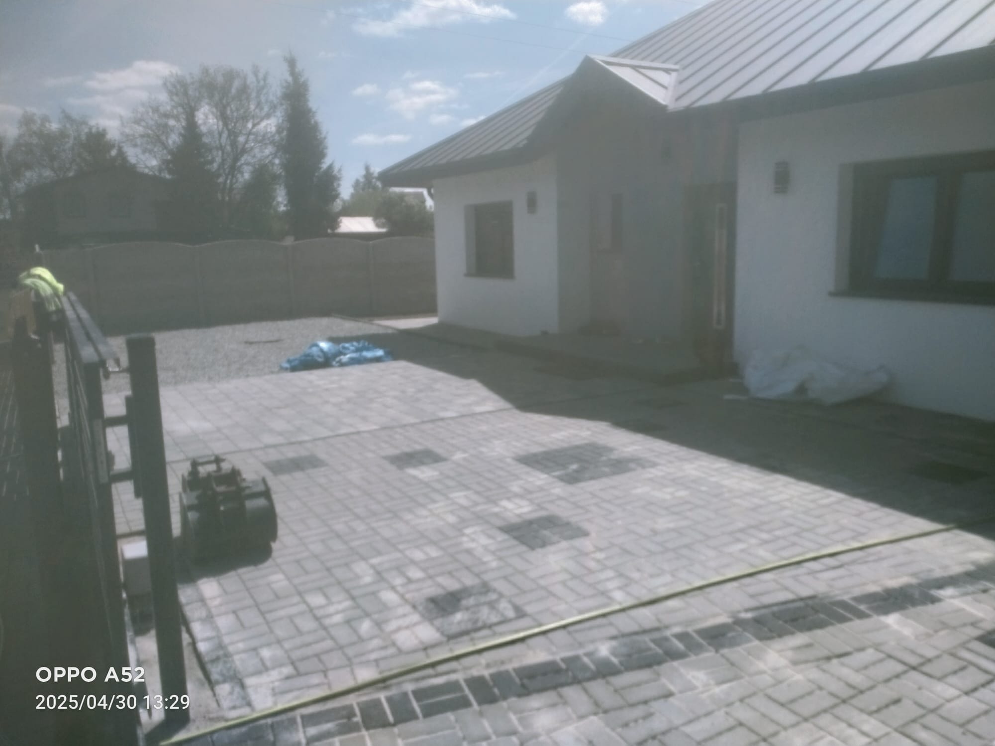 Ułożona szara kostka brukowa na podjeździe przed domem z szarą dachówką. Widoczny fragment ogrodzenia i sprzęt budowlany.