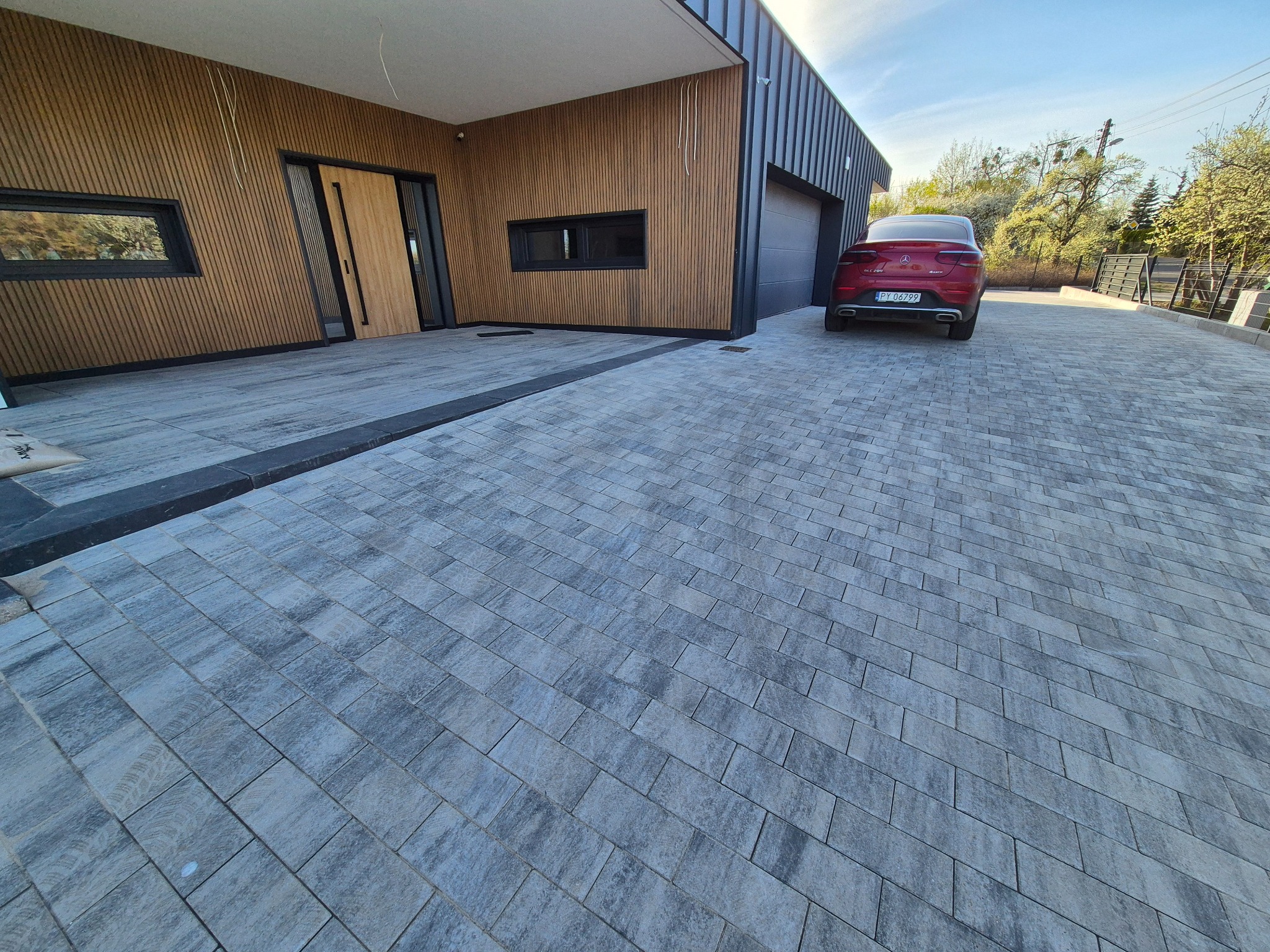 Nowoczesna posesja z szarej kostki brukowej ułożonej w jodełkę. Na podjeździe zaparkowany czerwony samochód. Budynek z drewnianą elewacją i czarnymi elementami.