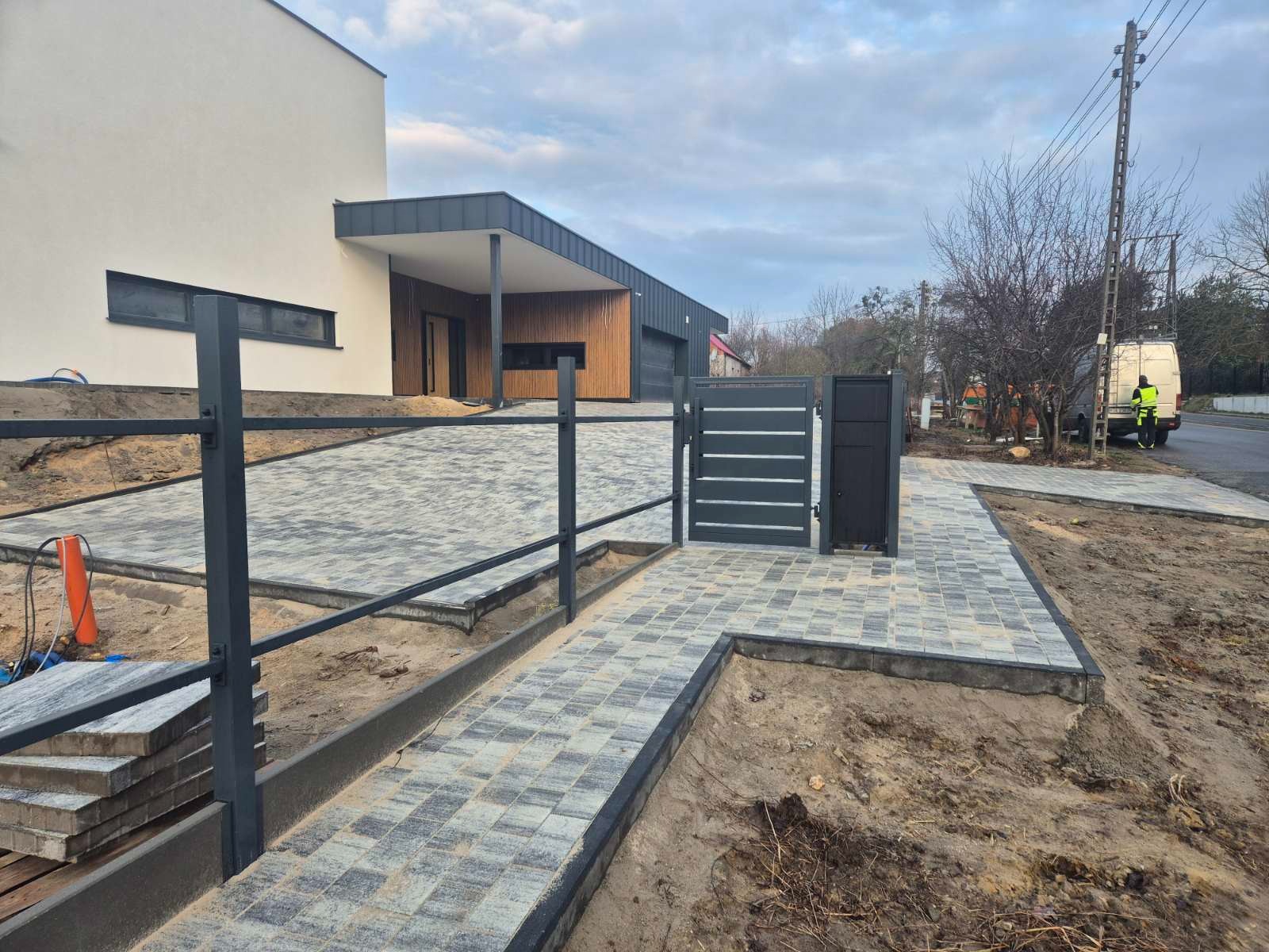 Nowoczesna posesja z szarą kostką brukową, metalowym ogrodzeniem i bramą. Widoczny fragment domu z drewnianą elewacją i podjazdem. Prace wykończeniowe w toku.