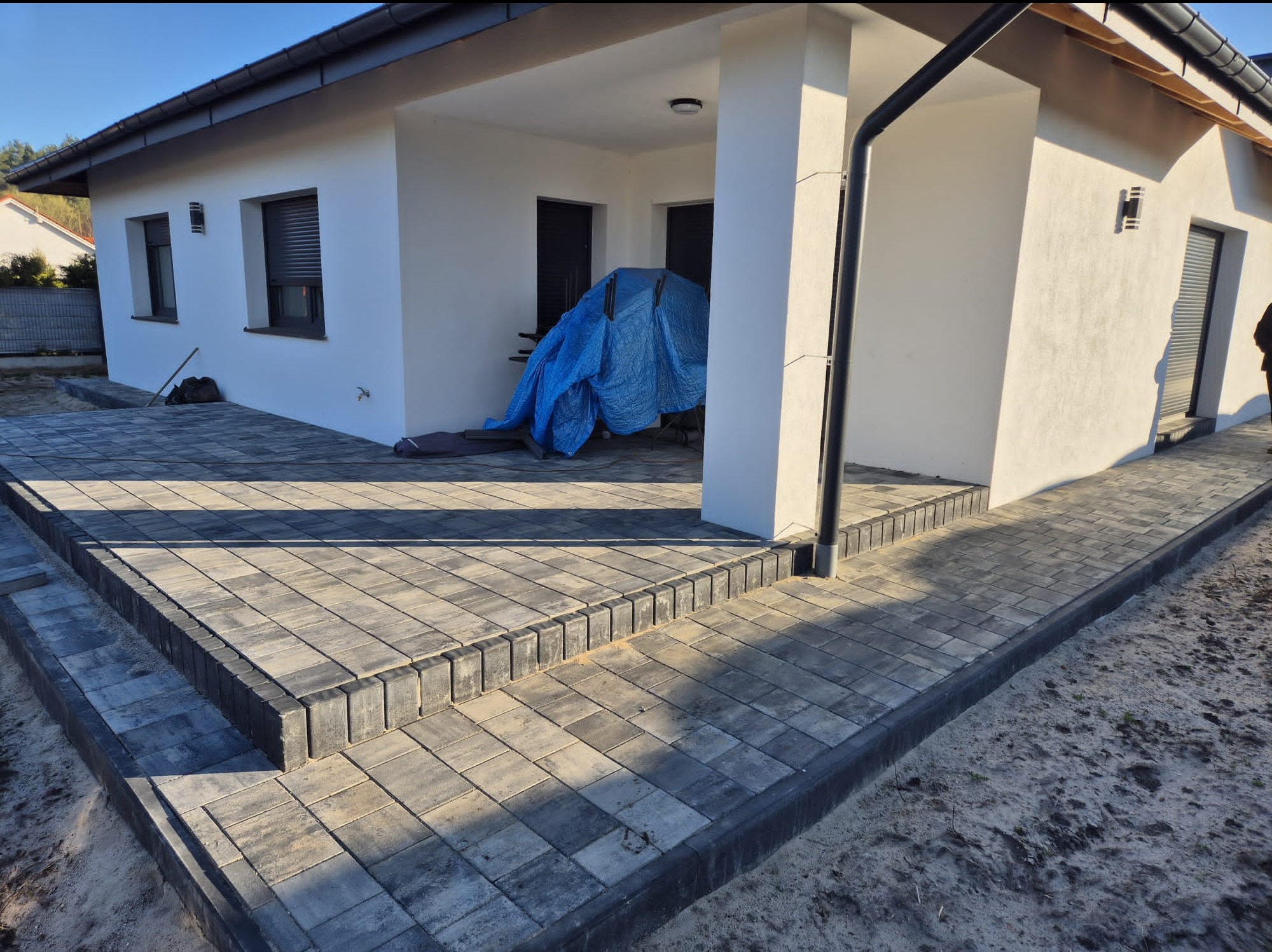 Szara kostka brukowa tworząca taras i ścieżkę przy białym domu. Ułożona z dbałością o detale, z widocznymi obrzeżami. Słoneczny dzień.