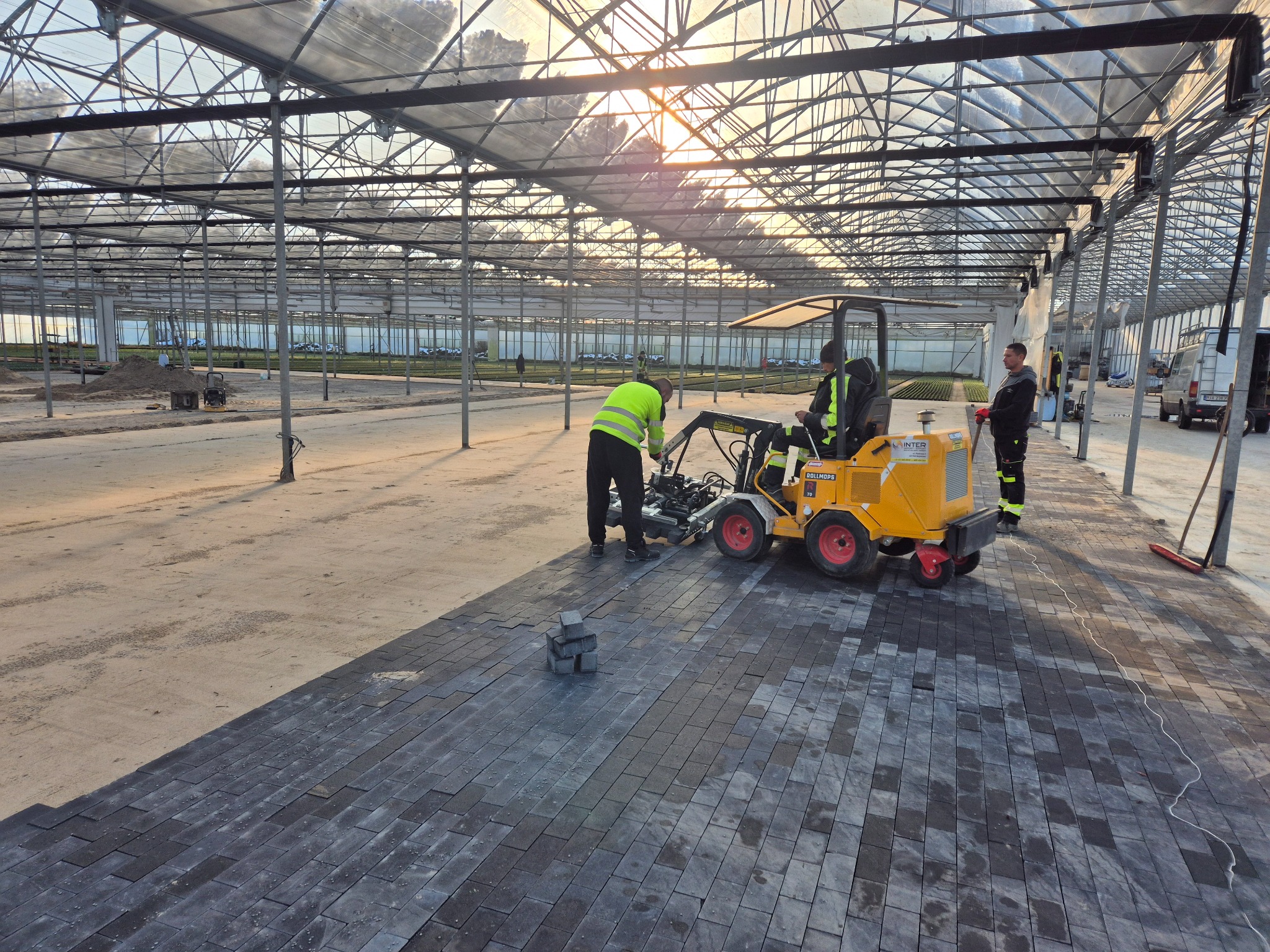 Układanie ciemnej kostki brukowej wewnątrz szklarni przy użyciu specjalistycznej maszyny. Widoczni pracownicy w odblaskowych kamizelkach i kostka brukowa.