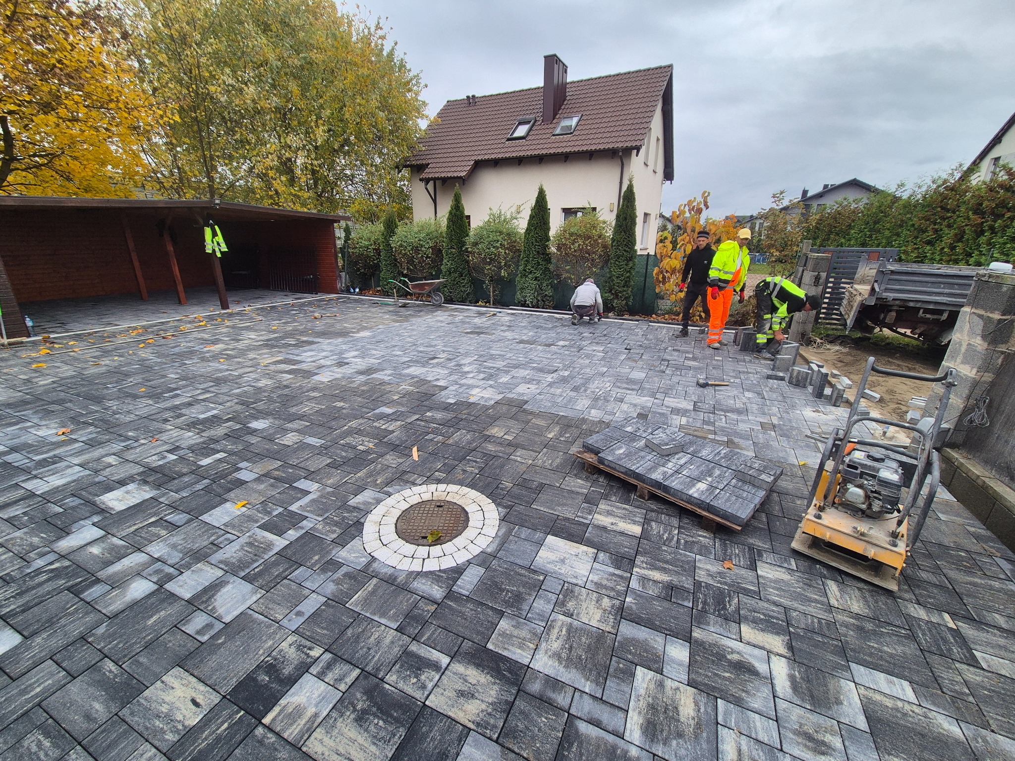 Układanie kostki brukowej na posesji z domem w tle. Widoczny fragment ułożonej nawierzchni, pracownicy w odblaskowych ubraniach i zagęszczarka wibracyjna.