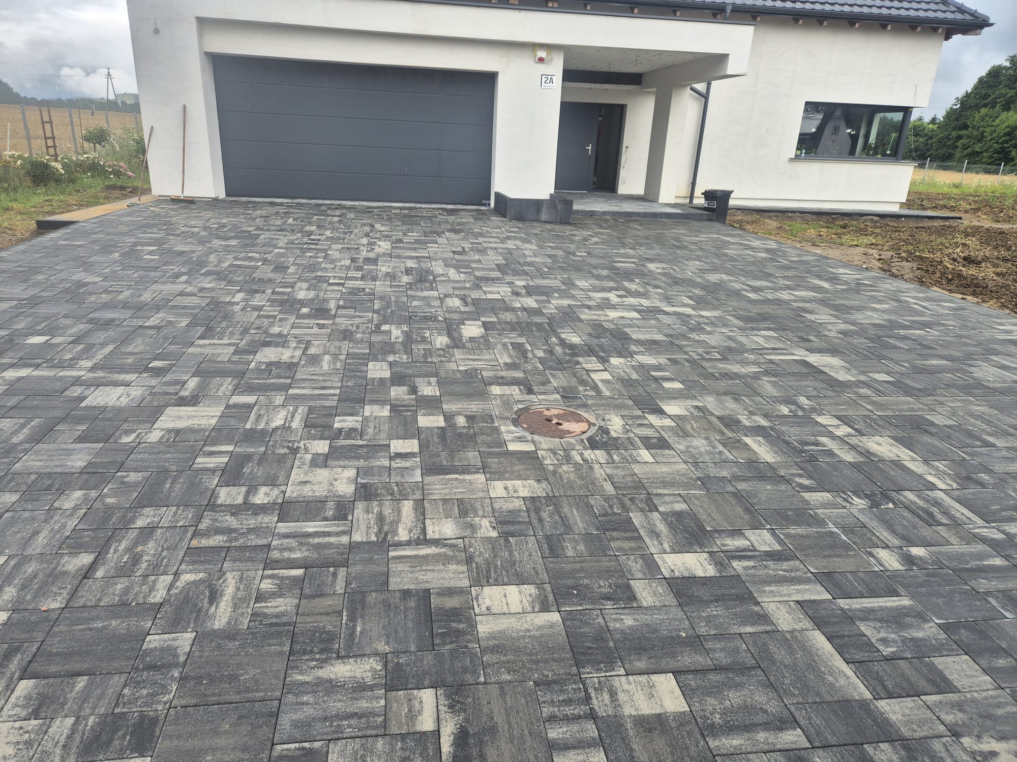 Nowoczesny podjazd z kostki brukowej w odcieniach szarości prowadzący do domu z garażem. Widoczny właz kanalizacyjny wkomponowany w nawierzchnię.