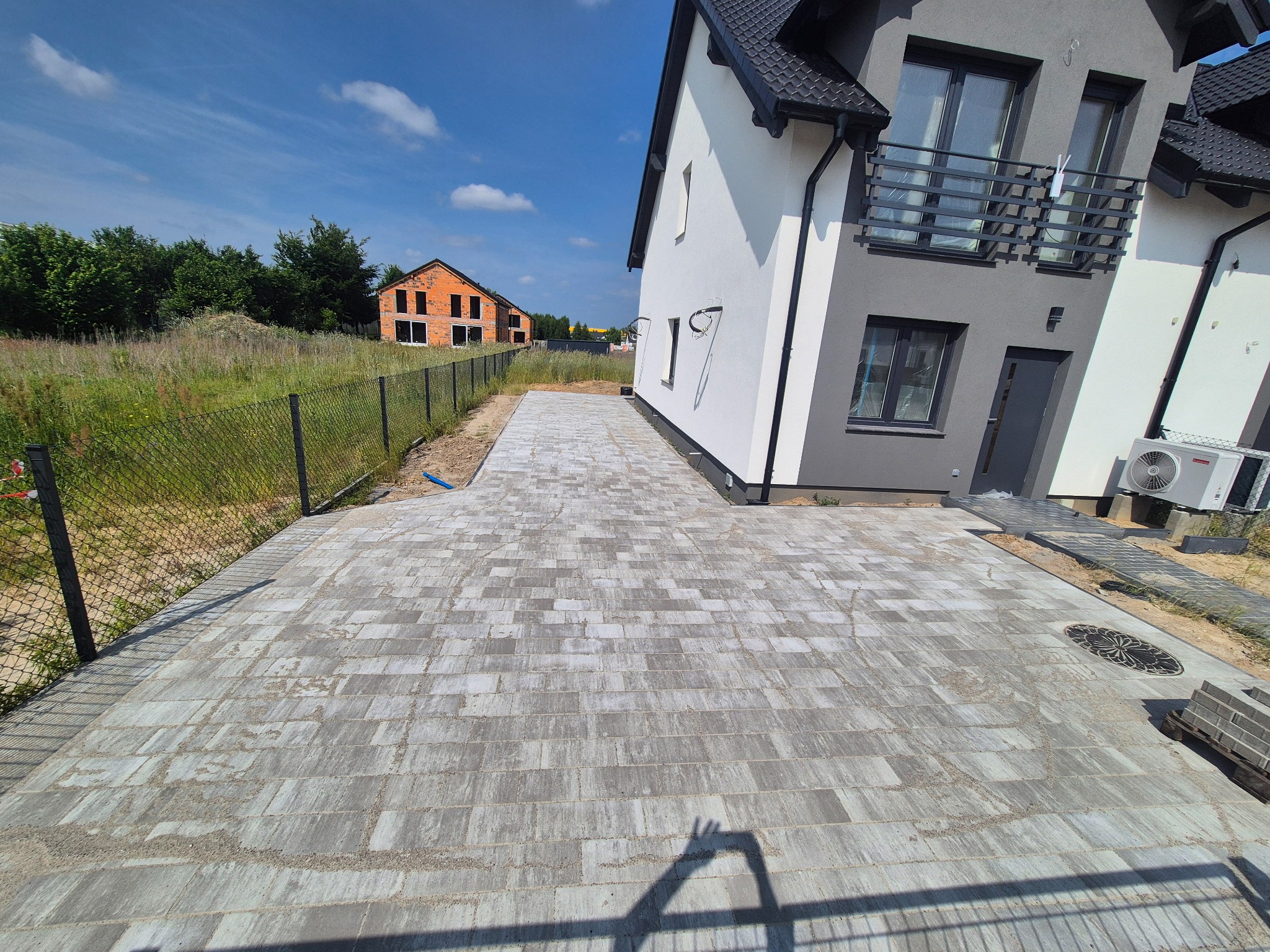 Ułożona szara kostka brukowa na podjeździe przed nowoczesnym domem z szarą elewacją. W tle budynek w stanie surowym i błękitne niebo.