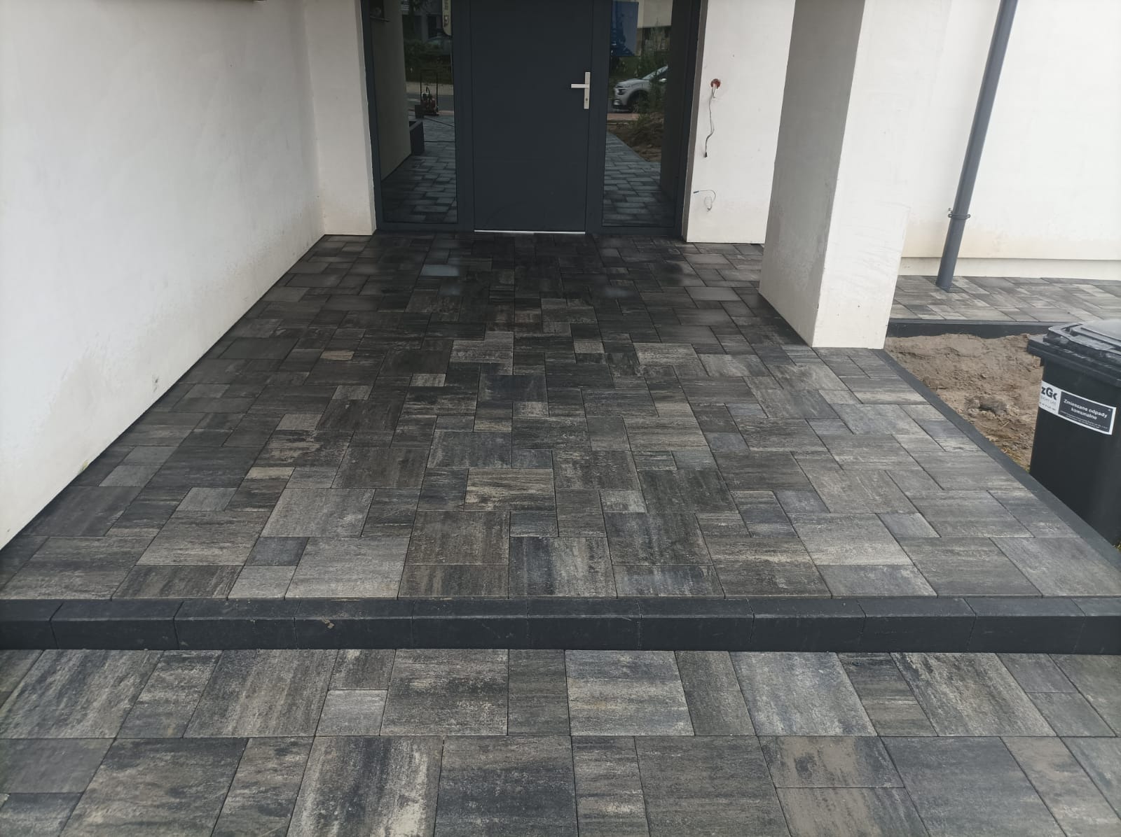 Nowoczesny podjazd z kostki brukowej w odcieniach szarości, z wyraźnym obramowaniem i schodkiem. Widoczny fragment elewacji budynku i drzwi wejściowe.