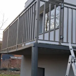 Nowoczesna balustrada balkonowa z metalowymi pionowymi szczeblami, widoczna z dołu, z drewnianą budą w tle i drabiną opartą o ścianę.