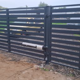 LEWSTAL PRZEMYSŁAW BABIŃSKI - Nowoczesna, automatyczna brama metalowa z poziomymi szczeblami, szara, zamontowana przy ogrodzeniu z siatki. Widoczne elementy automatyki bramy i podłączenia elektryczne.