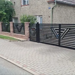 Nowoczesne ogrodzenie metalowe z bramą wjazdową, czarne poziome panele z motywem V, murowane słupki z cegły klinkierowej, kostka brukowa na podjeździe.