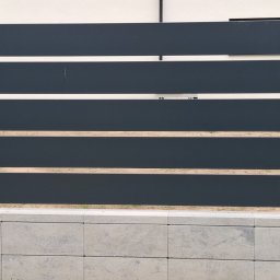 Lewstal - Nowoczesne ogrodzenie panelowe z ciemnoszarych, poziomych lameli metalowych, osadzone na słupkach i podmurówce z szarych bloczków betonowych.