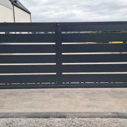 Lewstal - Nowoczesna, dwuskrzydłowa brama z poziomymi lamelami w kolorze antracytowym, osadzona między słupkami z szarych bloczków betonowych. Przed nią jasna kostka brukowa. Widoczny mechanizm.