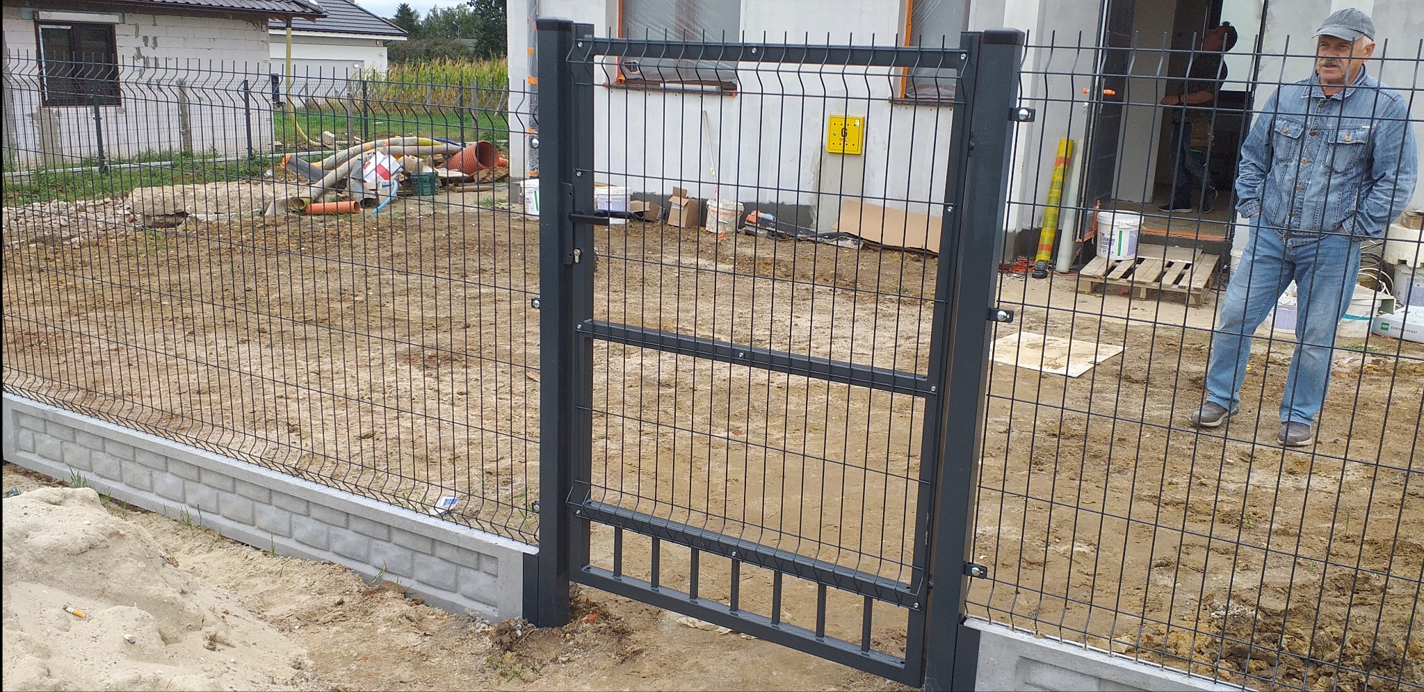 Ciemnoszara brama panelowa z siatki, zamontowana na betonowej podmurówce, z widocznym mężczyzną w dżinsowej kurtce w tle, na placu budowy.