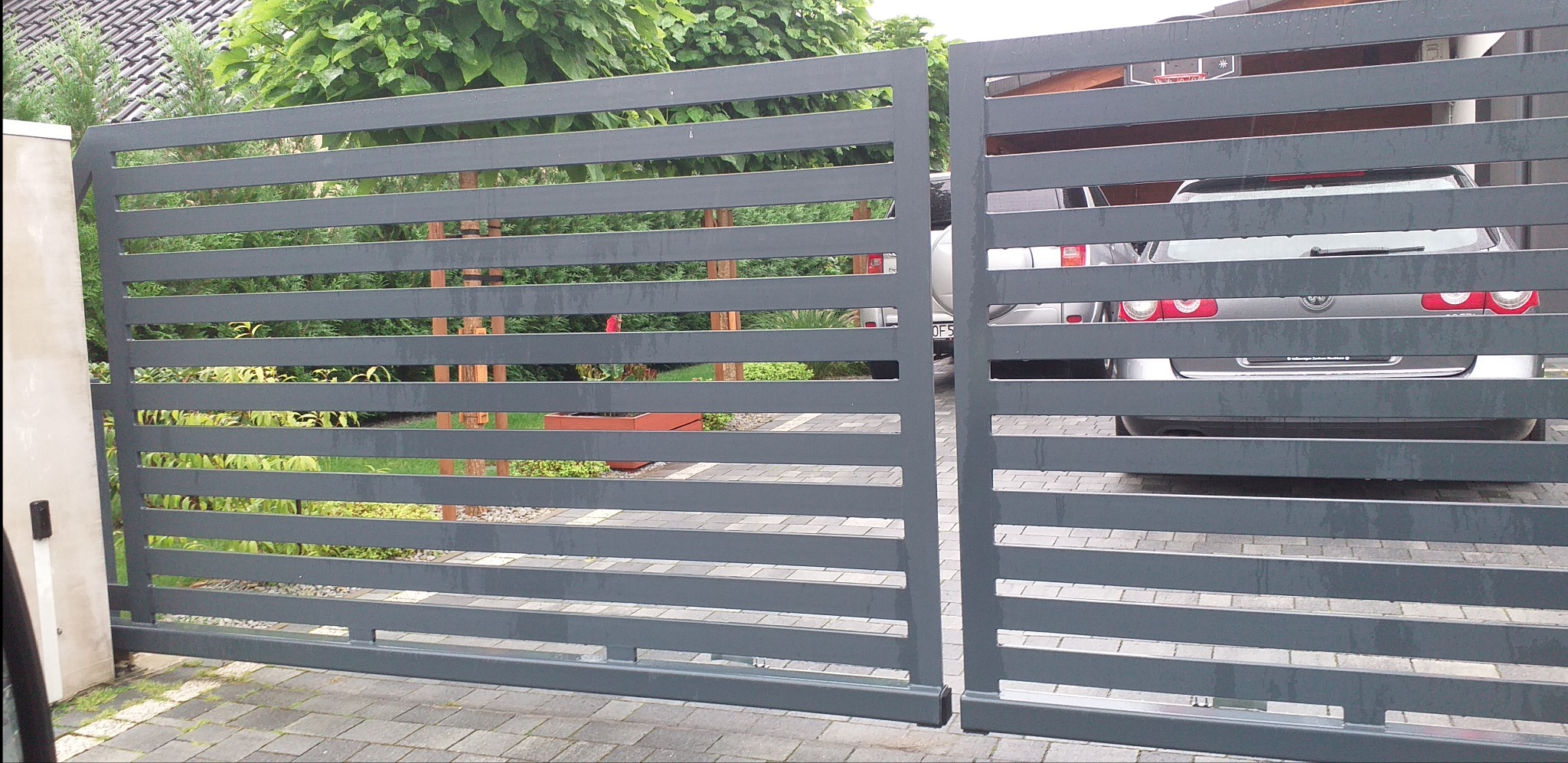 Otwarta, pozioma brama wjazdowa z szarych, metalowych profili, widok na zaparkowane samochody i zieleń ogrodu w tle.