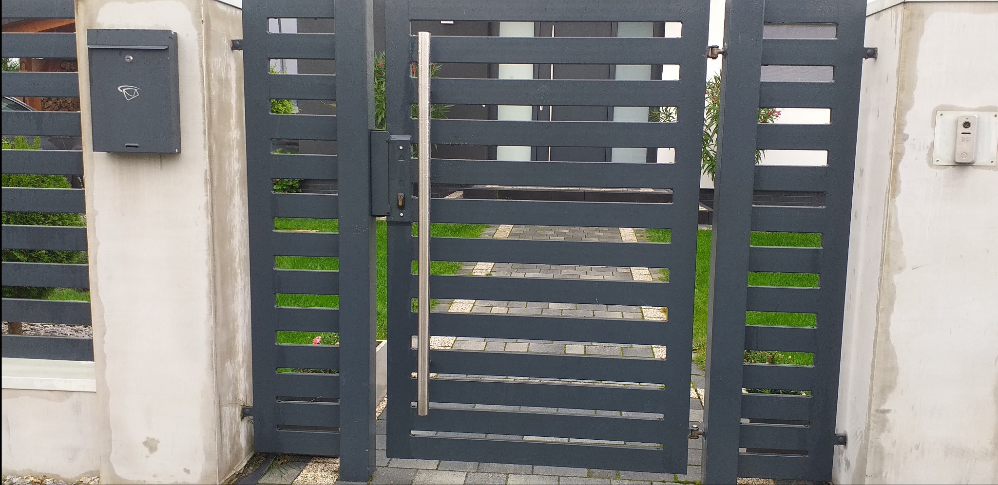 Nowoczesna, antracytowa brama wjazdowa z poziomymi panelami, z długim, pionowym uchwytem ze stali nierdzewnej, zamontowana między betonowymi słupkami z domofonem i skrzynką na listy, widok...