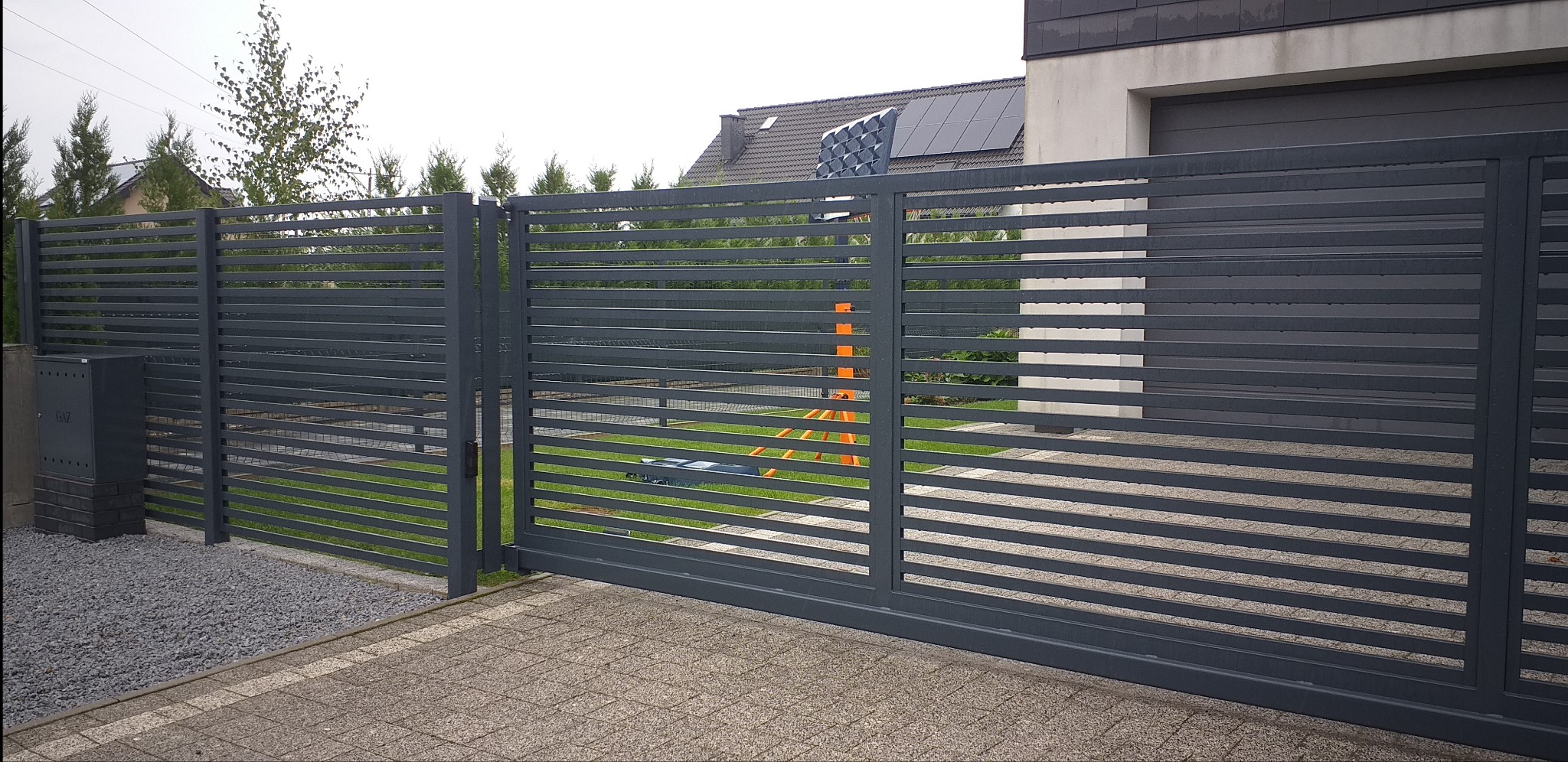 Nowoczesna, pozioma, metalowa brama przesuwna w Olszance, z widocznym w tle domem z panelami słonecznymi, ustawiona na kostce brukowej, z fragmentem ogrodzenia.