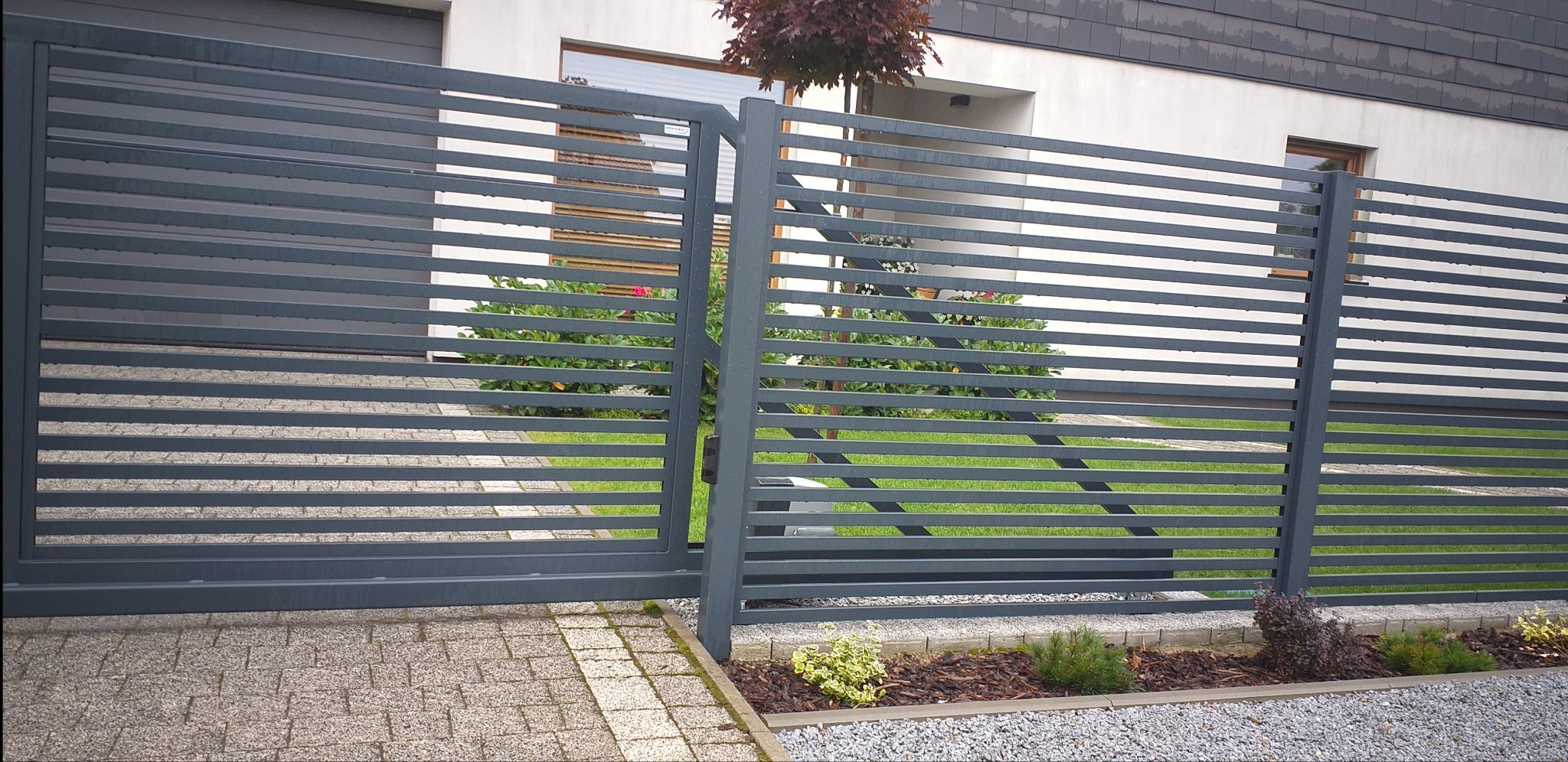 Nowoczesna, przesuwna brama wjazdowa z poziomych, metalowych profili w kolorze grafitowym, z widocznym mechanizmem automatycznego otwierania, na tle domu z szarą dachówką i starannie utrzymanym...