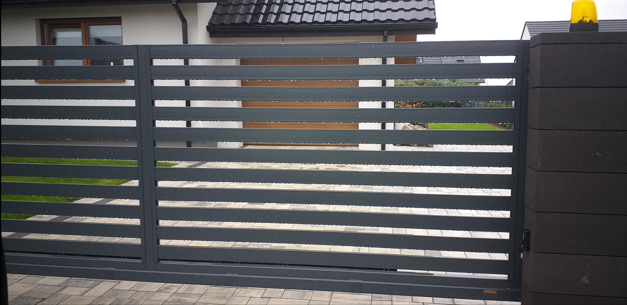 Nowoczesna, przesuwna brama wjazdowa z poziomych, grafitowych paneli metalowych, z żółtą lampą ostrzegawczą na słupku z szarej kostki, widok z zewnątrz na posesję z kostką brukową i trawnikiem.