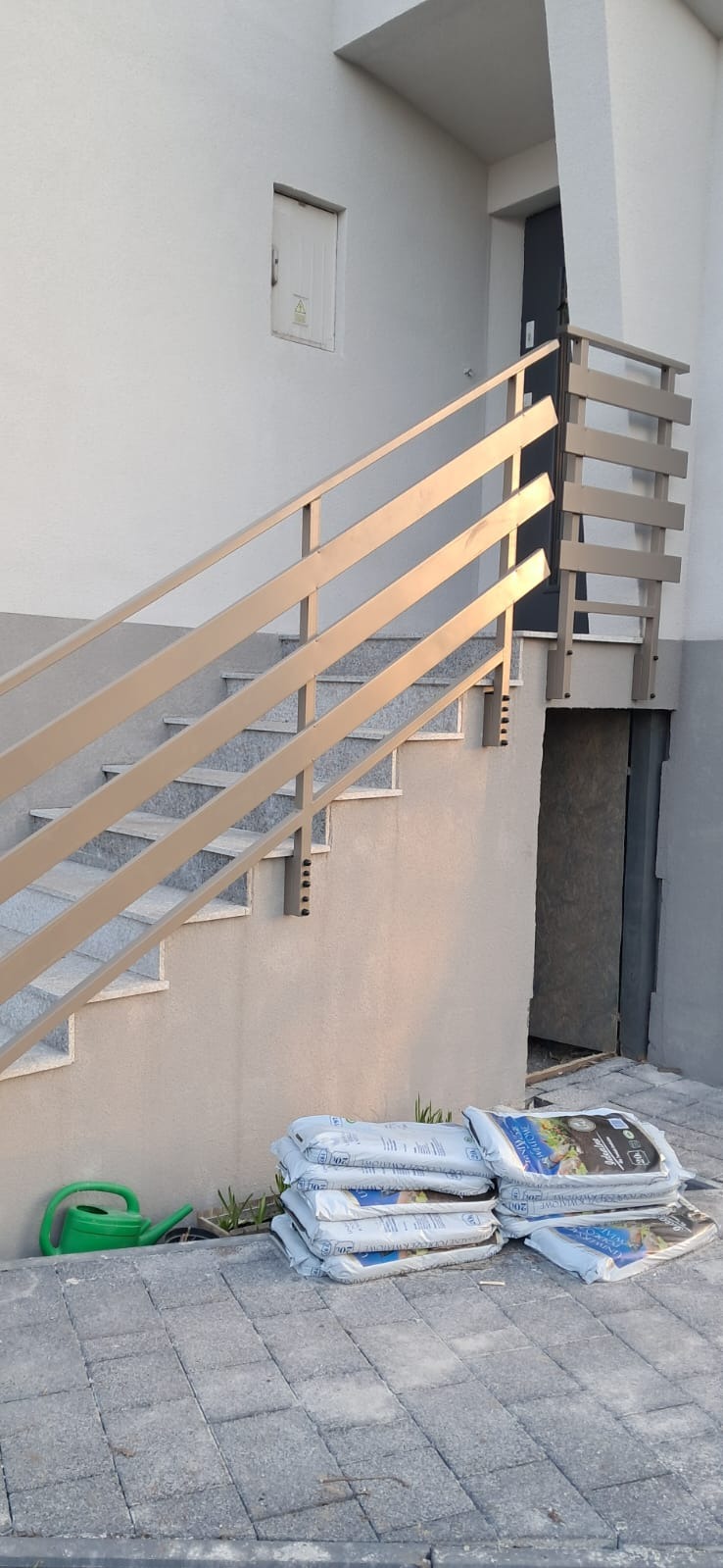 Zewnętrzne schody z metalową balustradą w kolorze beżowym, ułożone worki ziemi i zielona konewka u podnóża schodów.