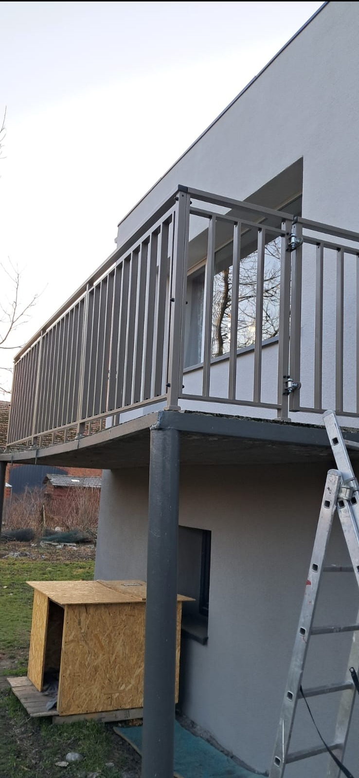 Nowoczesna balustrada balkonowa z metalowymi pionowymi szczeblami, widoczna z dołu, z drewnianą budą w tle i drabiną opartą o ścianę.