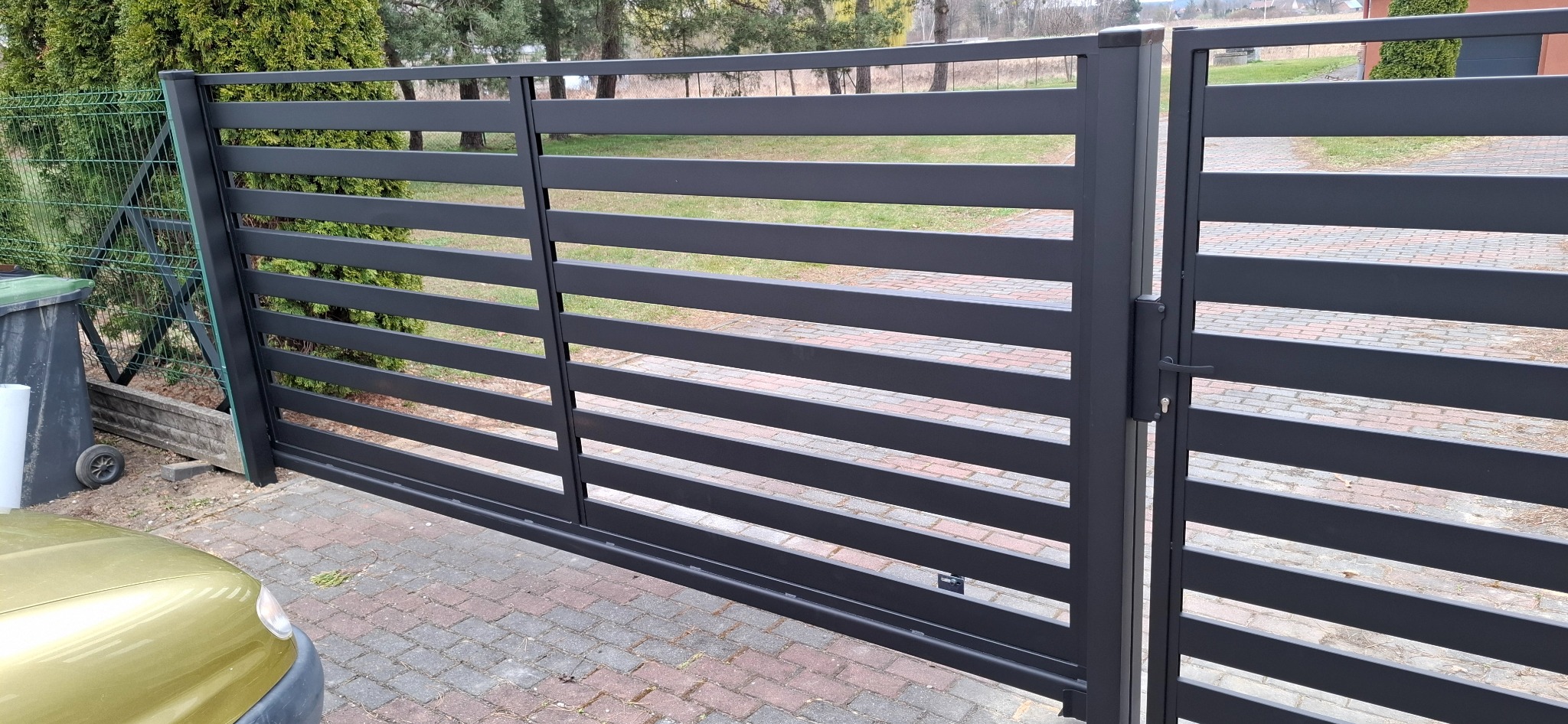 Nowoczesna, przesuwna brama wjazdowa z poziomymi, metalowymi panelami w kolorze grafitowym, widoczna na tle kostki brukowej i zieleni ogrodu.