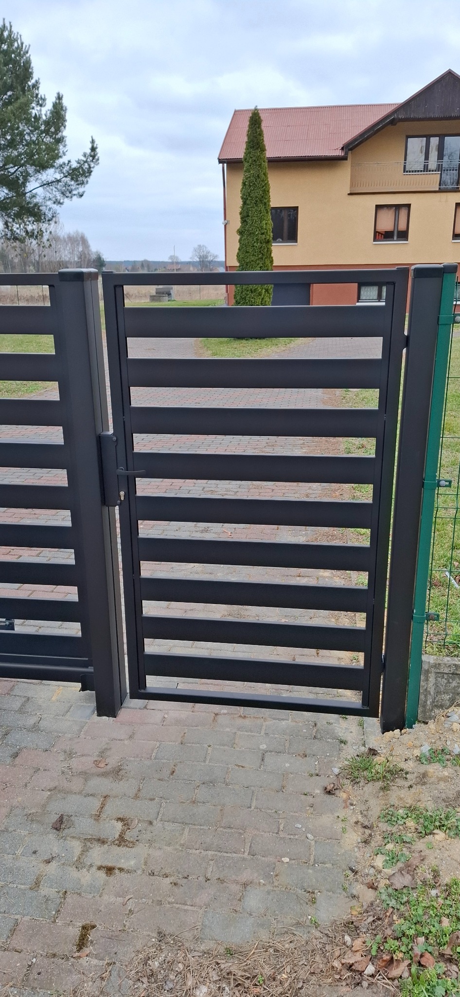 Nowoczesna, pozioma, czarna brama wjazdowa z zamkiem, widok z zewnątrz na tle domu z brązowym dachem i zielonym drzewem.