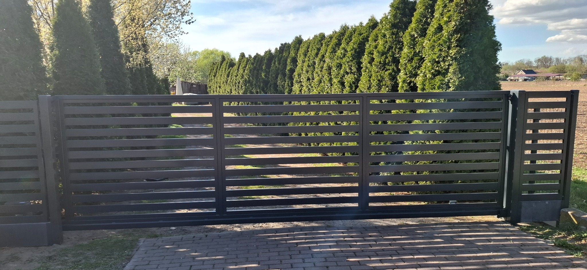 Nowoczesna, przesuwna brama wjazdowa z poziomych, metalowych profili, z widocznymi cieniami na brukowanej nawierzchni podjazdu i szpalerem wysokich tuj w tle.
