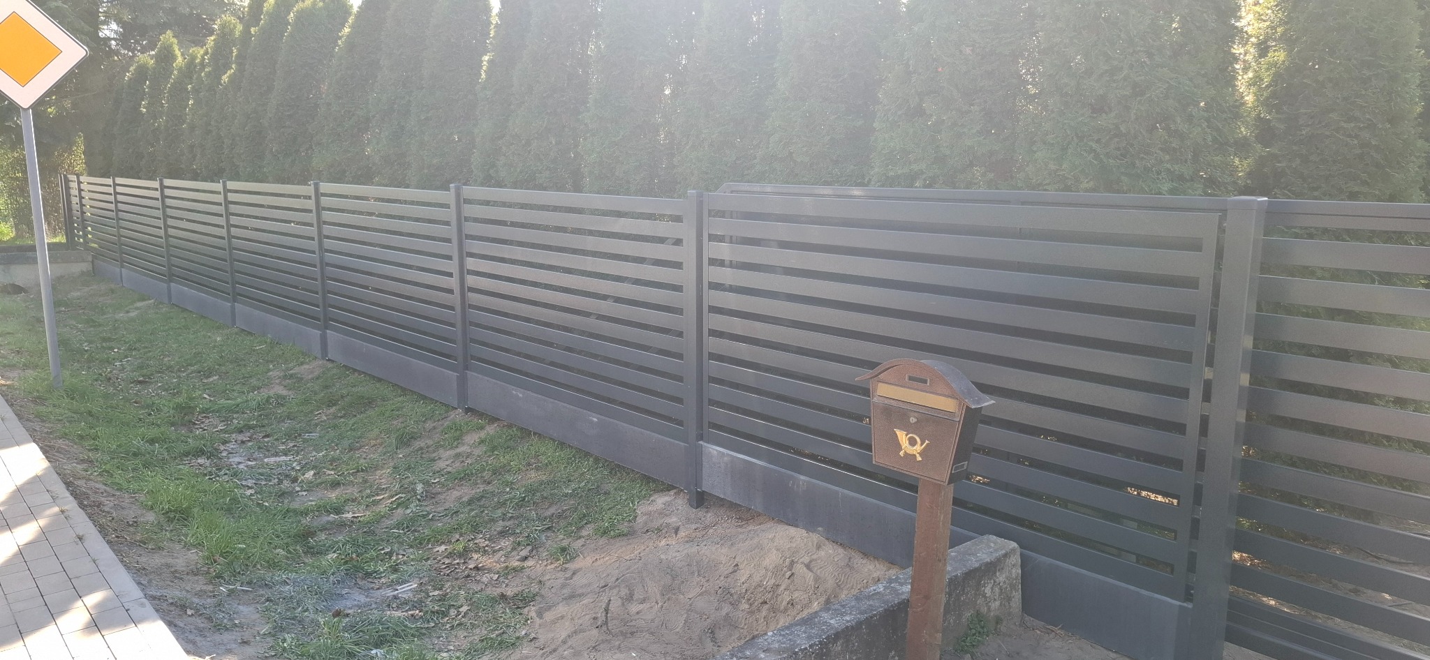 Nowoczesne, poziome przęsła w szarym kolorze, tworzące ogrodzenie posesji, z widoczną skrzynką pocztową w stylu retro i znakiem drogowym w tle.